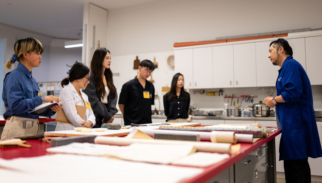 Five people listen to an instructor in a blue coat in a bright workshop, discussing scholarships, with materials on large tables.