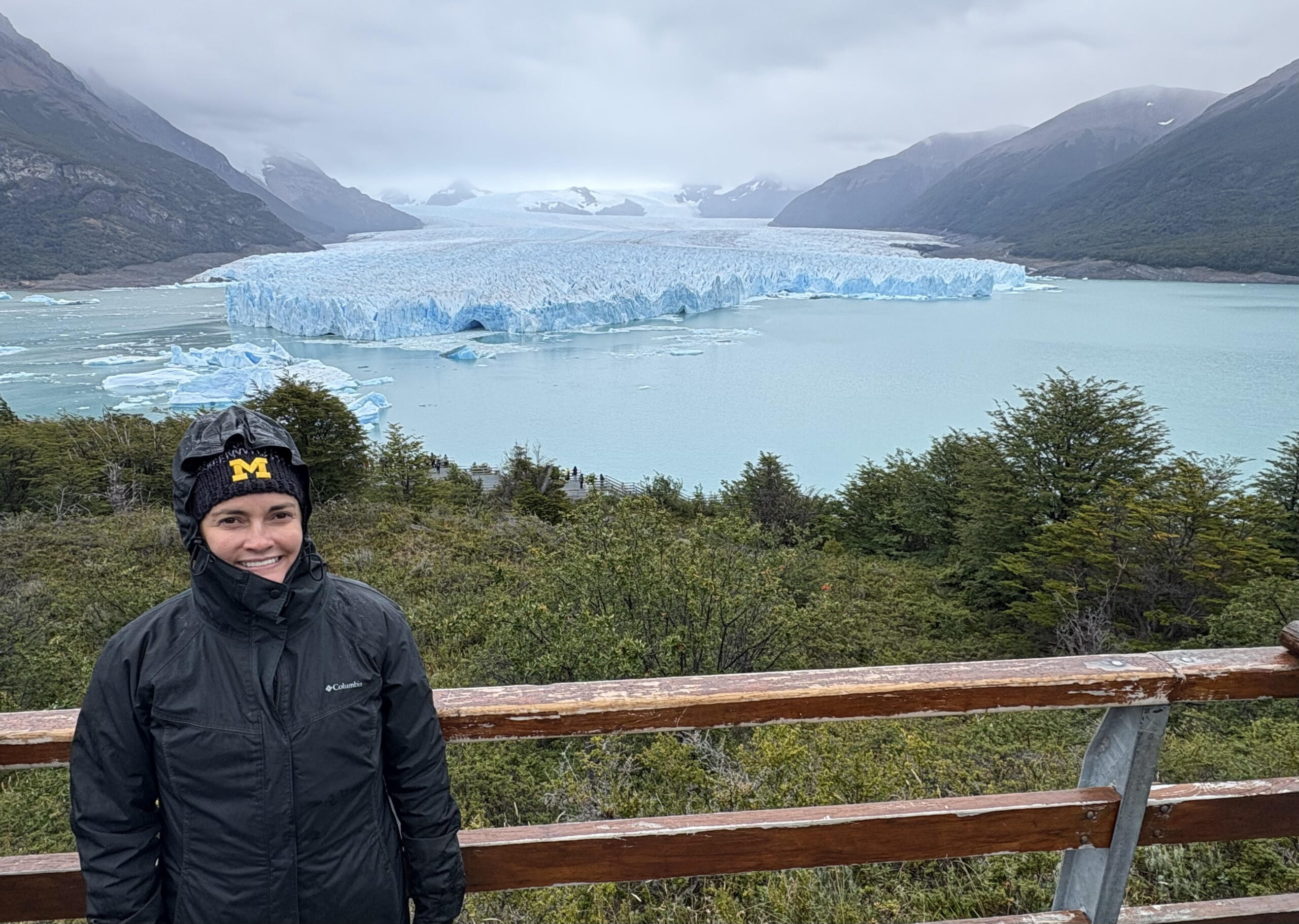 A person in a black jacket and hat stands by a wooden railing, smiling, with a blue glacier, turquoise water, and mountains behind.