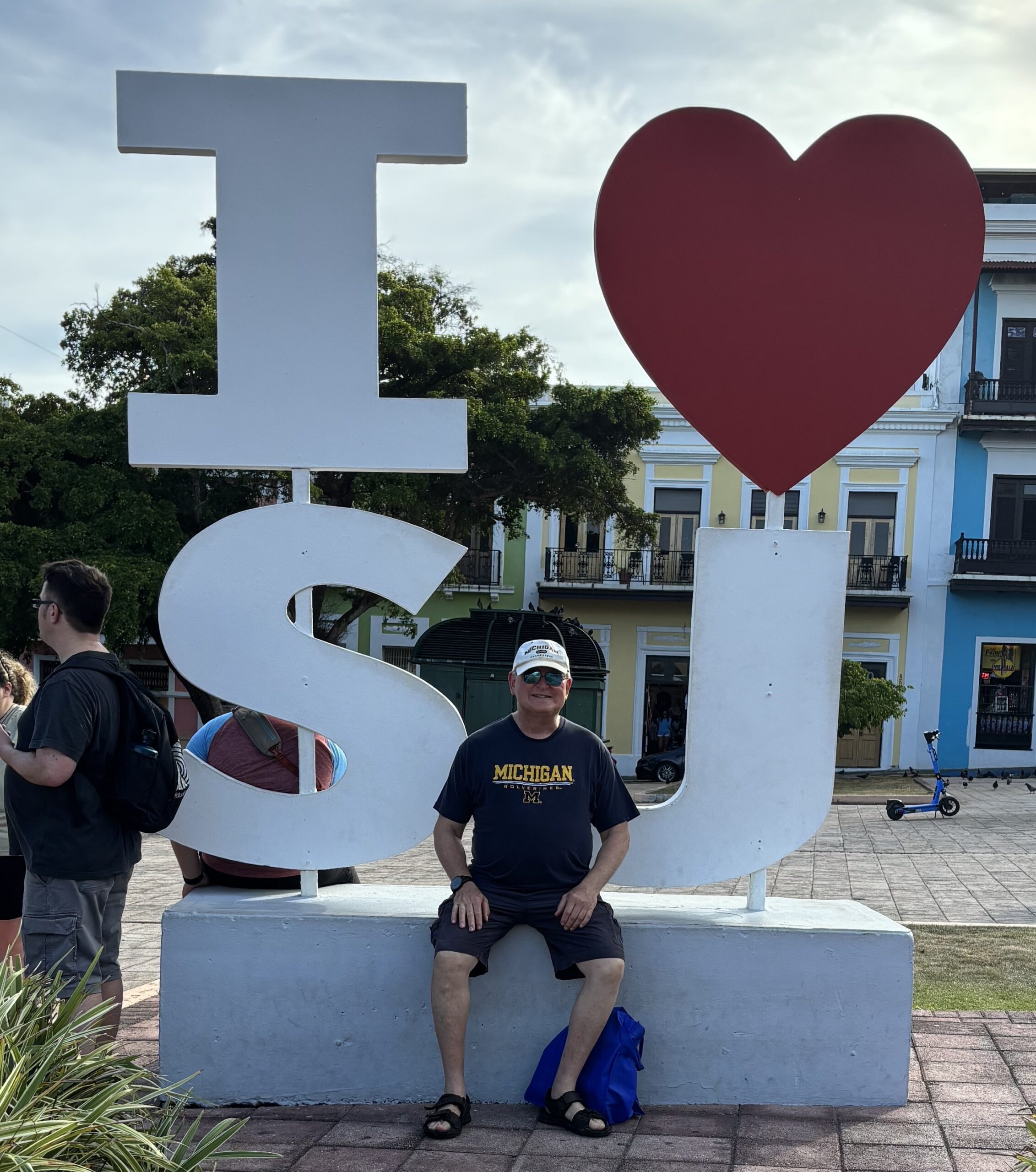 A man in a blue shirt sits on a bench before a large I ♥ SJ sign in a busy square, amid colorful buildings, and crowds.