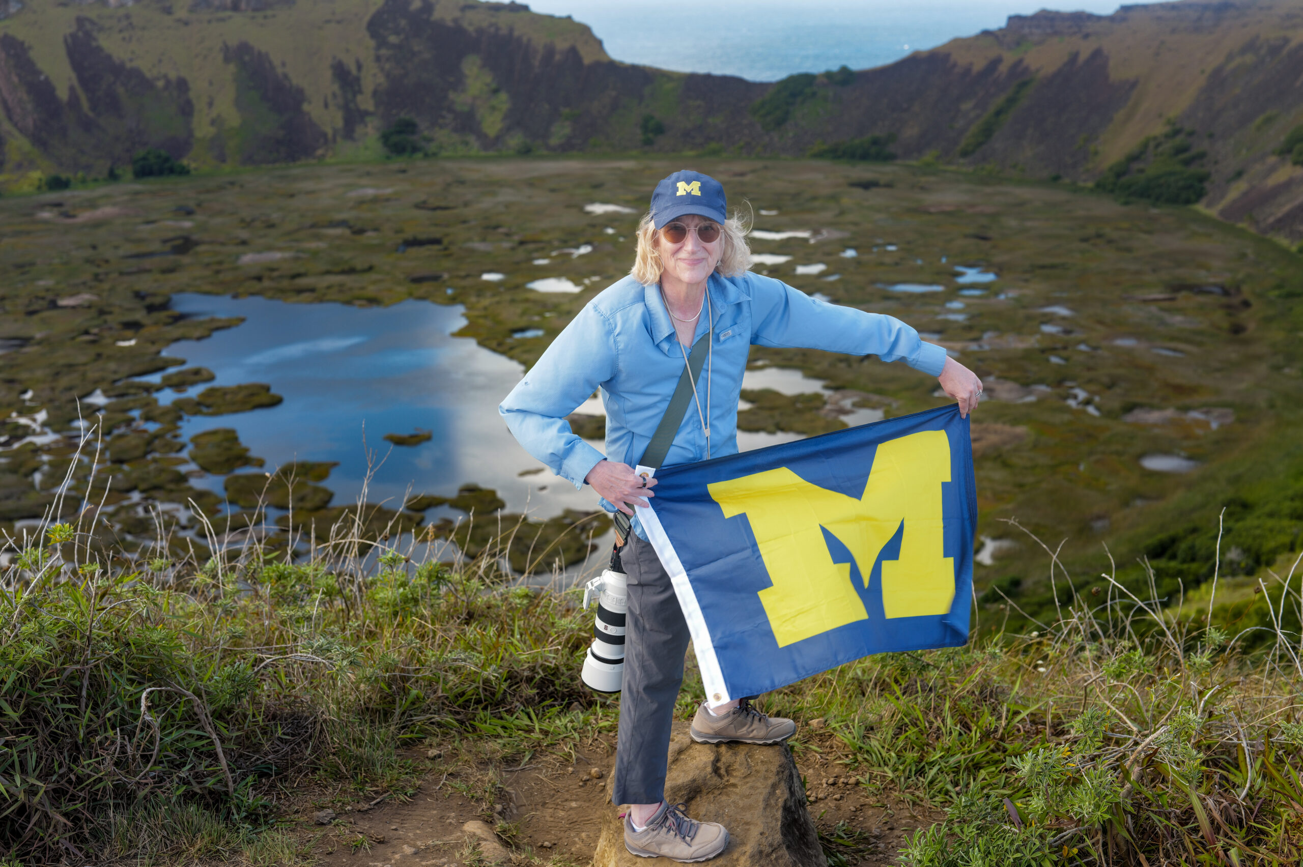 A person in a blue shirt and cap with a camera holds a University of Michigan flag on a hill, overlooking a crater with water and grass.