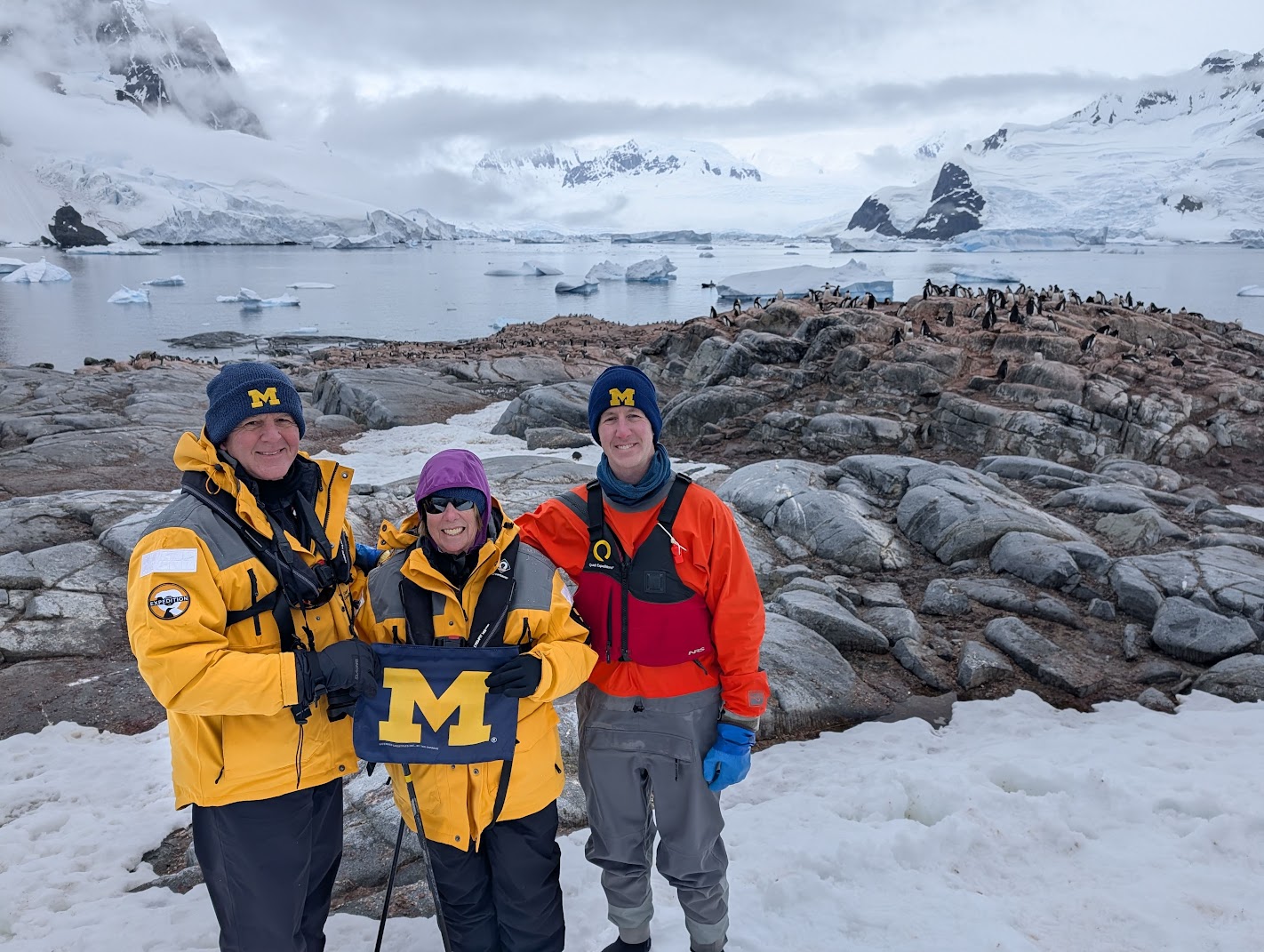 Three people in blue and gold winter gear stand on Antarctic rocks, holding a Michigan M flag. Penguins, icy mountains complete the scene.