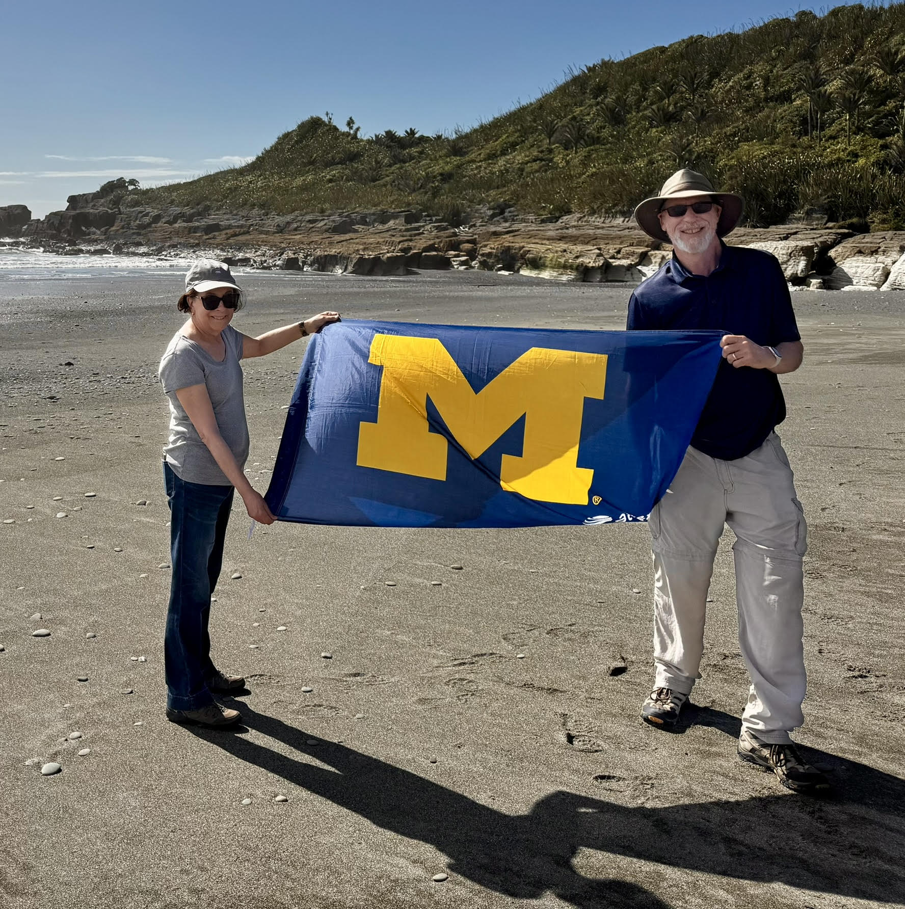 Two people stand on a sandy beach, holding a blue flag with a yellow M. Green hills, trees, and the ocean shine under a clear sky.