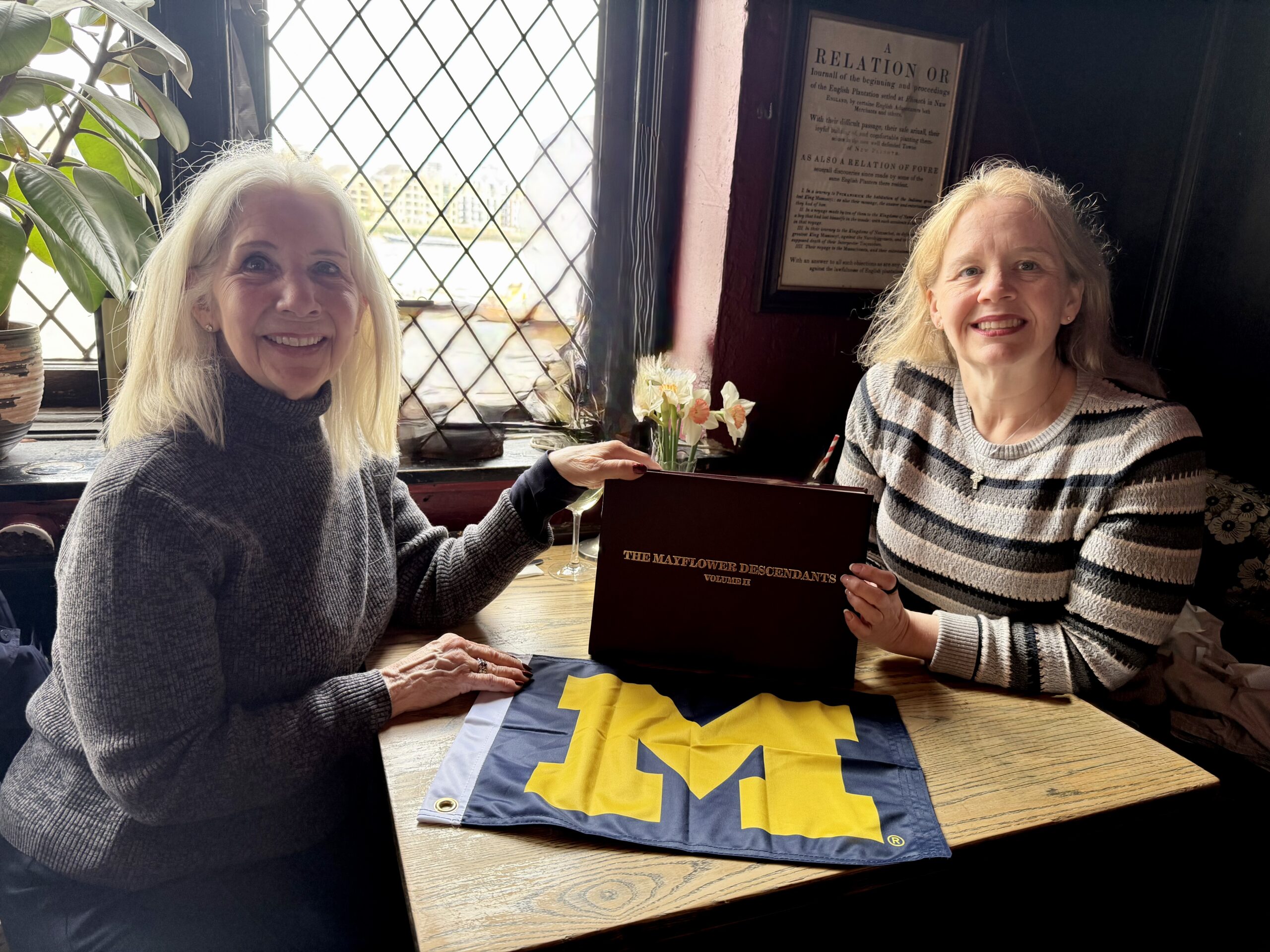 Two women at a wooden table by a window smile at the camera with a Mayflower Descendants folder, and a Michigan flag on the table.