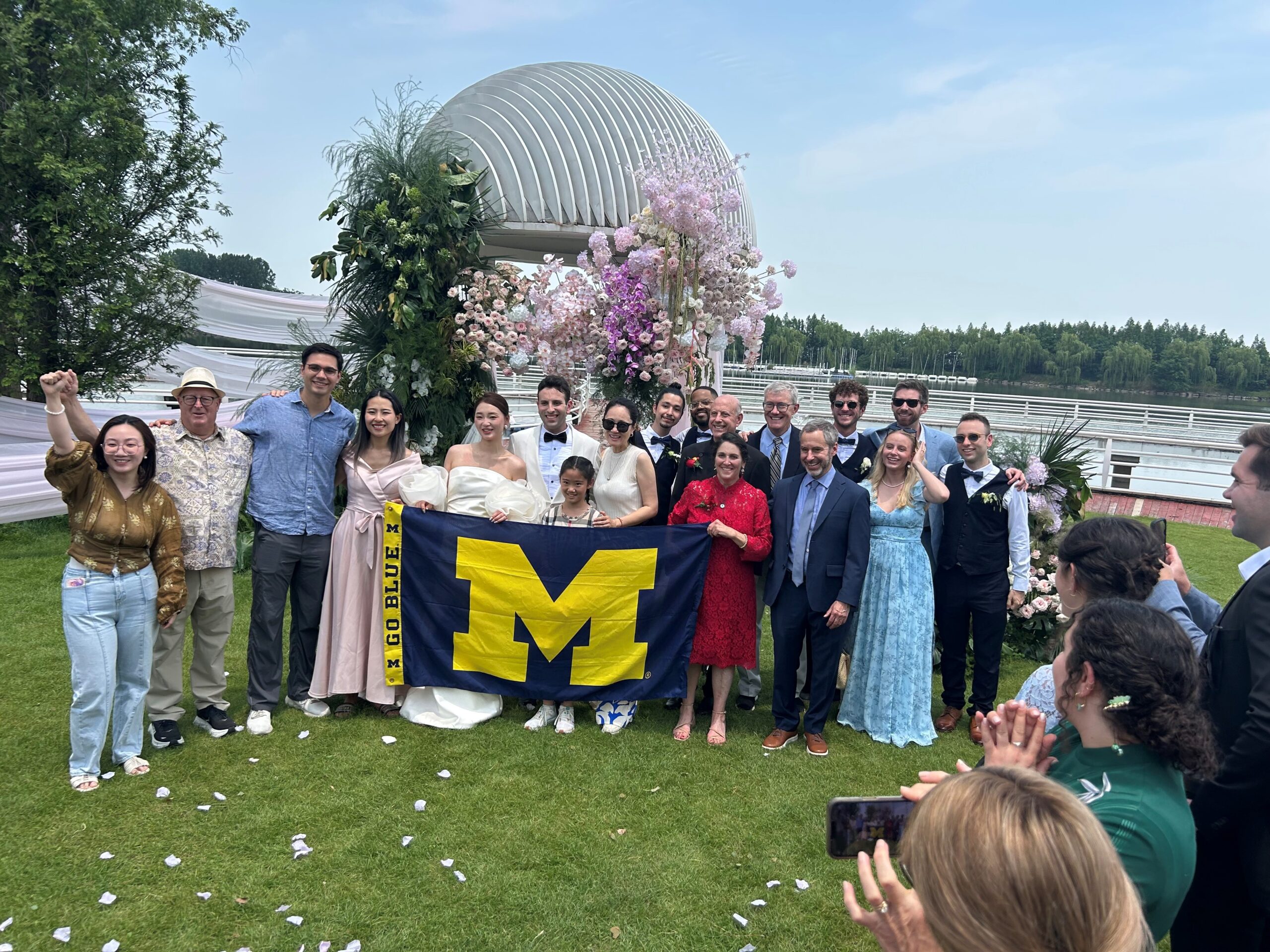 A group poses at a wedding, holding a Michigan Blue flag before a pink-and-white floral arch, as guests clap and cheer.