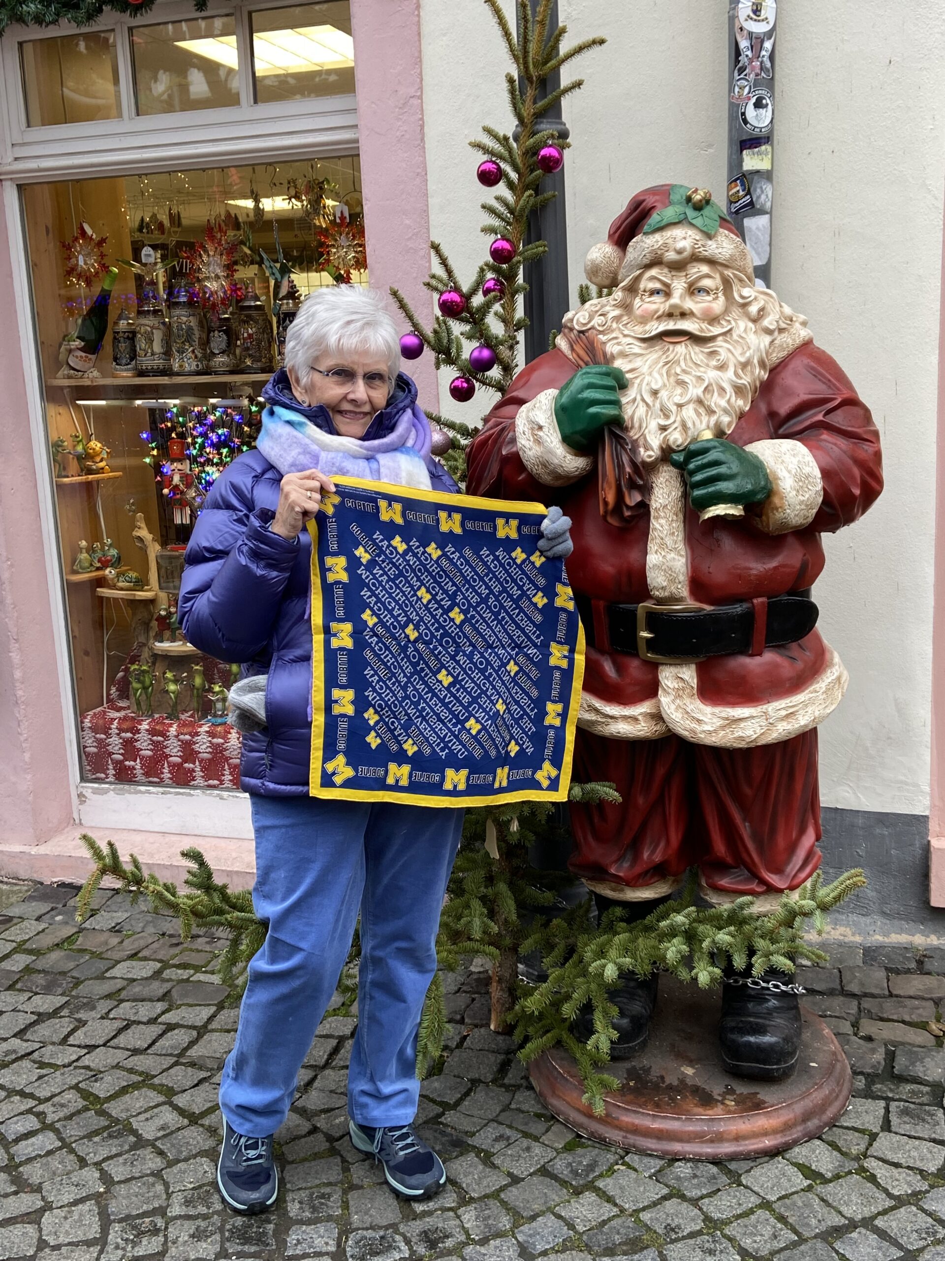 An older woman in a purple jacket stands next to a Santa Claus statue outdoors, holding a blue and yellow Michigan cloth, with festive decorations, and World cheer in the background.