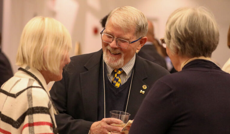 A older man with glasses and a beard wearing a suit talks to two women.