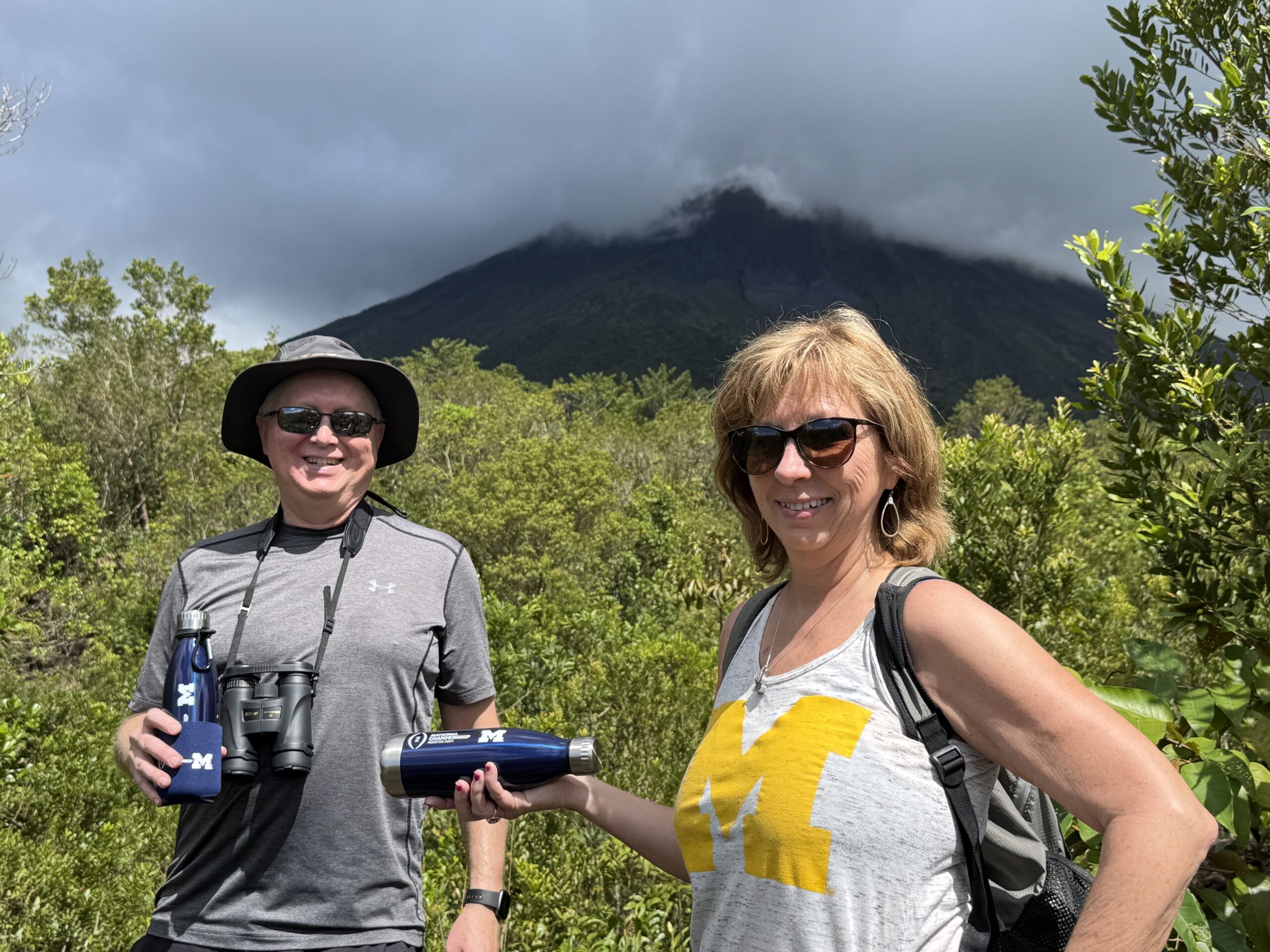A man and woman in sunglasses smile while hiking before a lush, green forest and cloud-covered volcano, holding Michigan “M” bottles.
