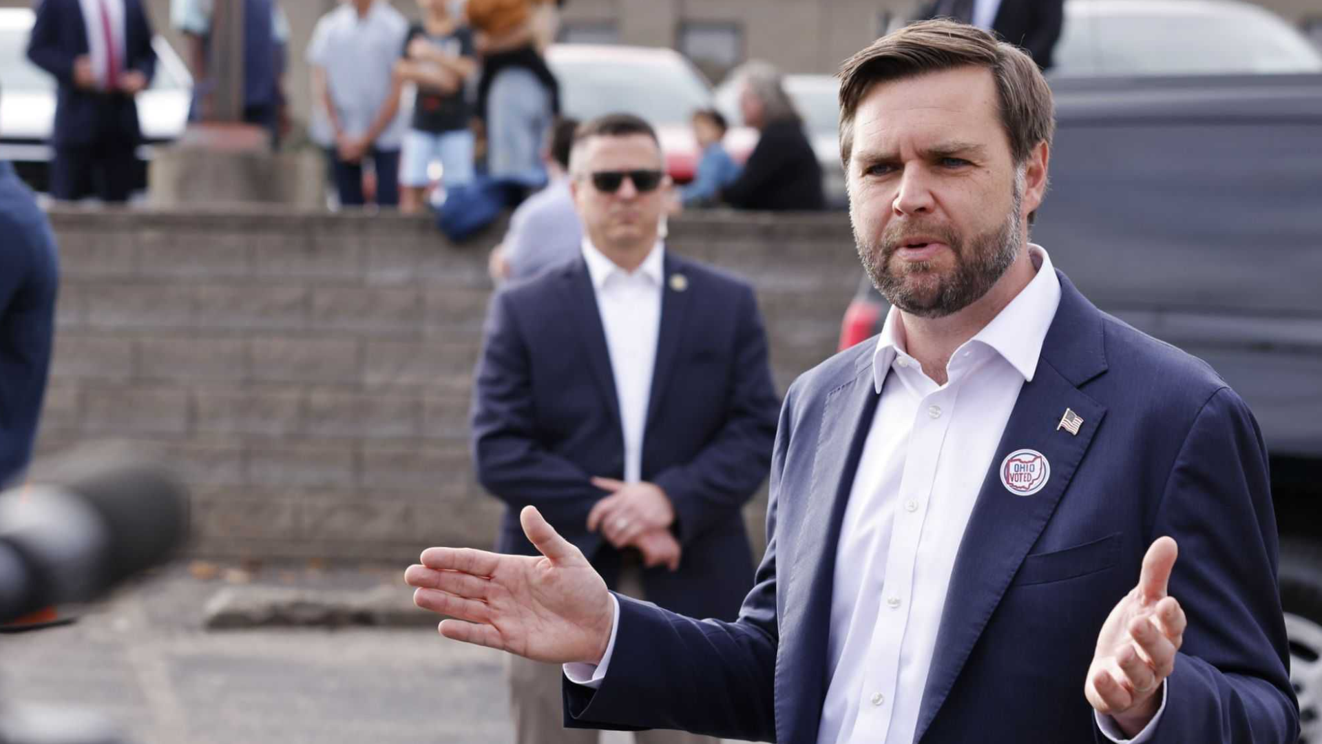 Matt Cybert, in a suit, stands behind JD Vance speaking to a group.