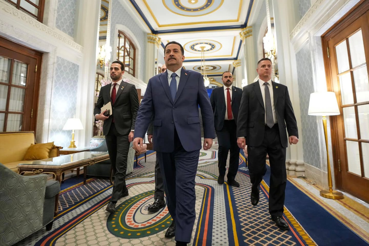 A group of men in suits walk towards the camera in an ornate hallway.