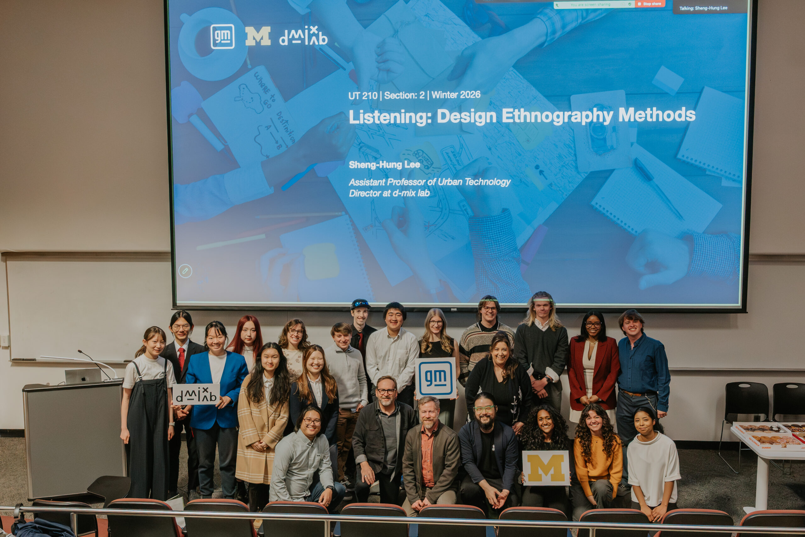 A diverse group of students and faculty pose in a classroom before a screen reading “Listening: Design Ethnography Methods,” some holding signs with logos and the University of Michigan “M.”.