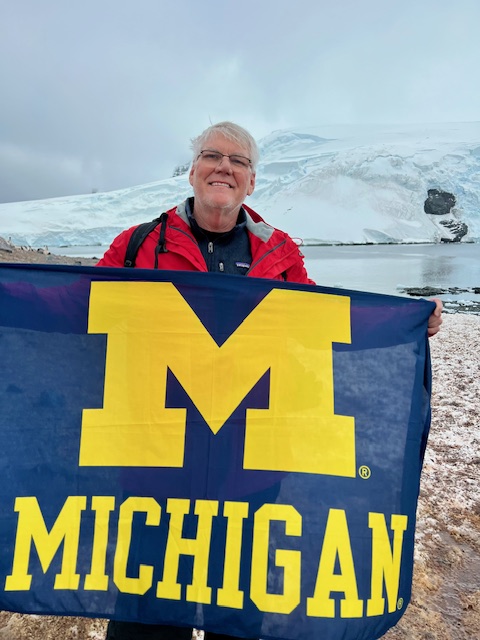 A smiling person in a blue and red jacket holds a blue and yellow University of Michigan flag in a snowy, icy landscape with mountains, water.