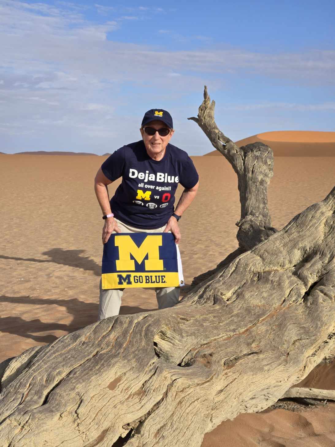 A person in a University of Michigan shirt and hat stands in a desert, holding a GO BLUE banner beside a weathered tree.