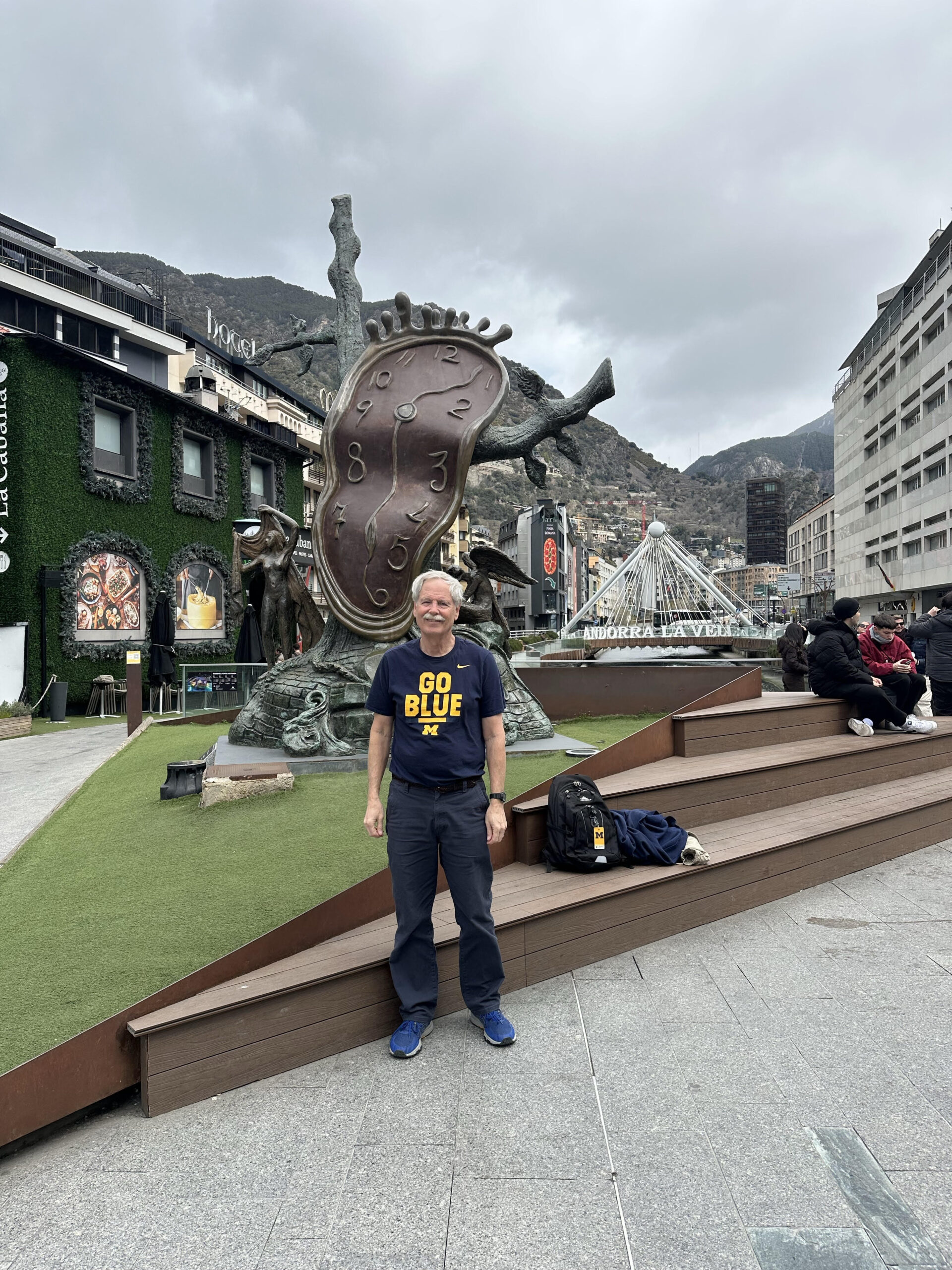 A man in a Go Blue T-shirt smiles by a Dalí melting clock in a city square, with mountains, buildings, and clouds behind him.