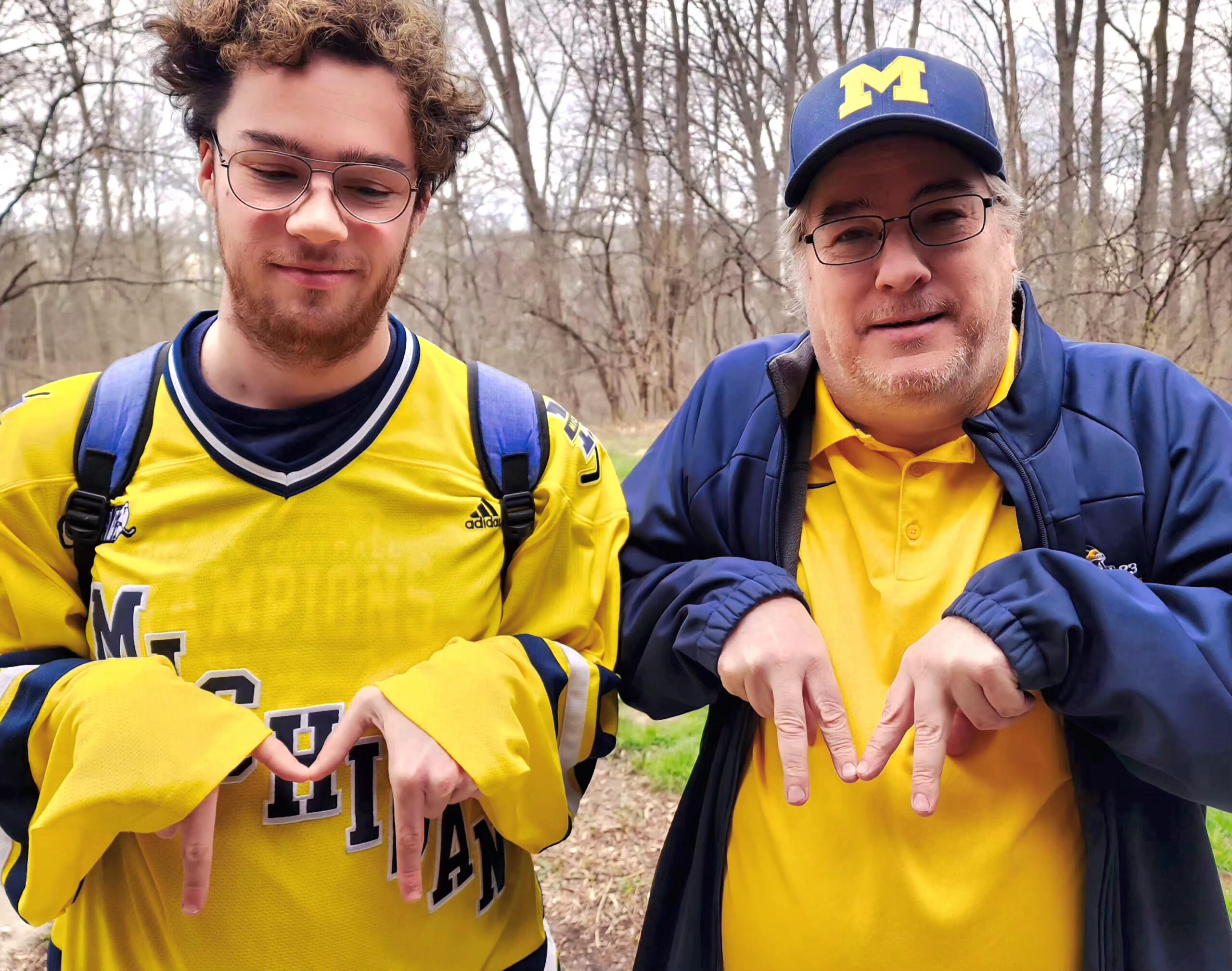 Two people in University of Michigan gear stand outside in blue, smiling and making the M sign, trees behind them.