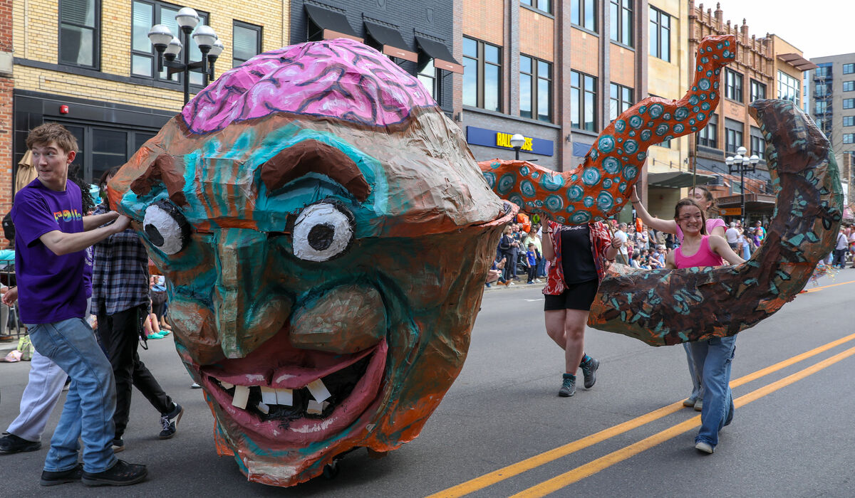 Students march down a downtown Ann Arbor Street with a giant paper mache head of a green creature with purple hair.