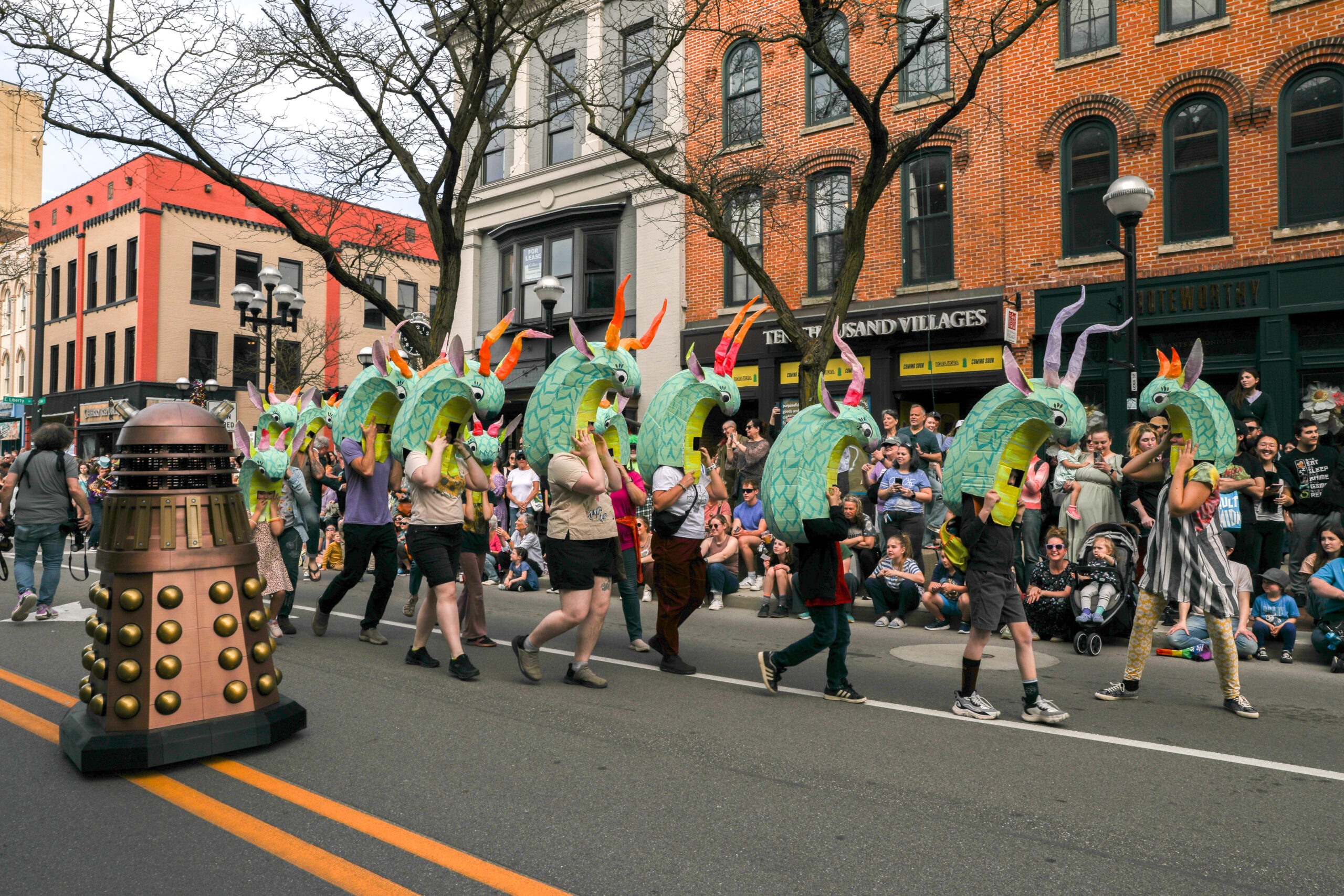 People in colorful dragon or serpent headpieces march in a city parade, with a Dalek nearby, as spectators watch and take photos.