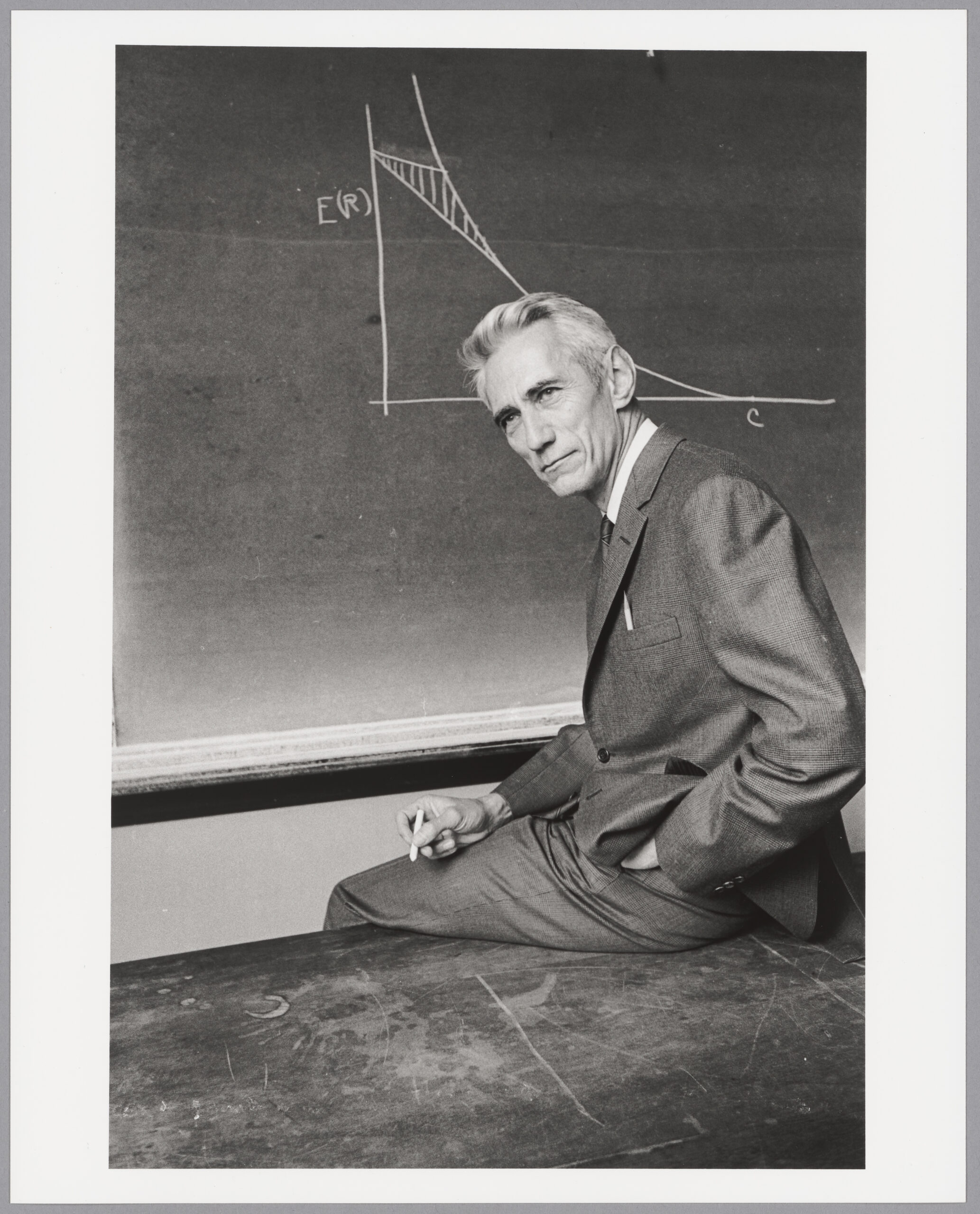 Claude Shannon sits at a desk facing a chalkboard with a chart drawn on it.