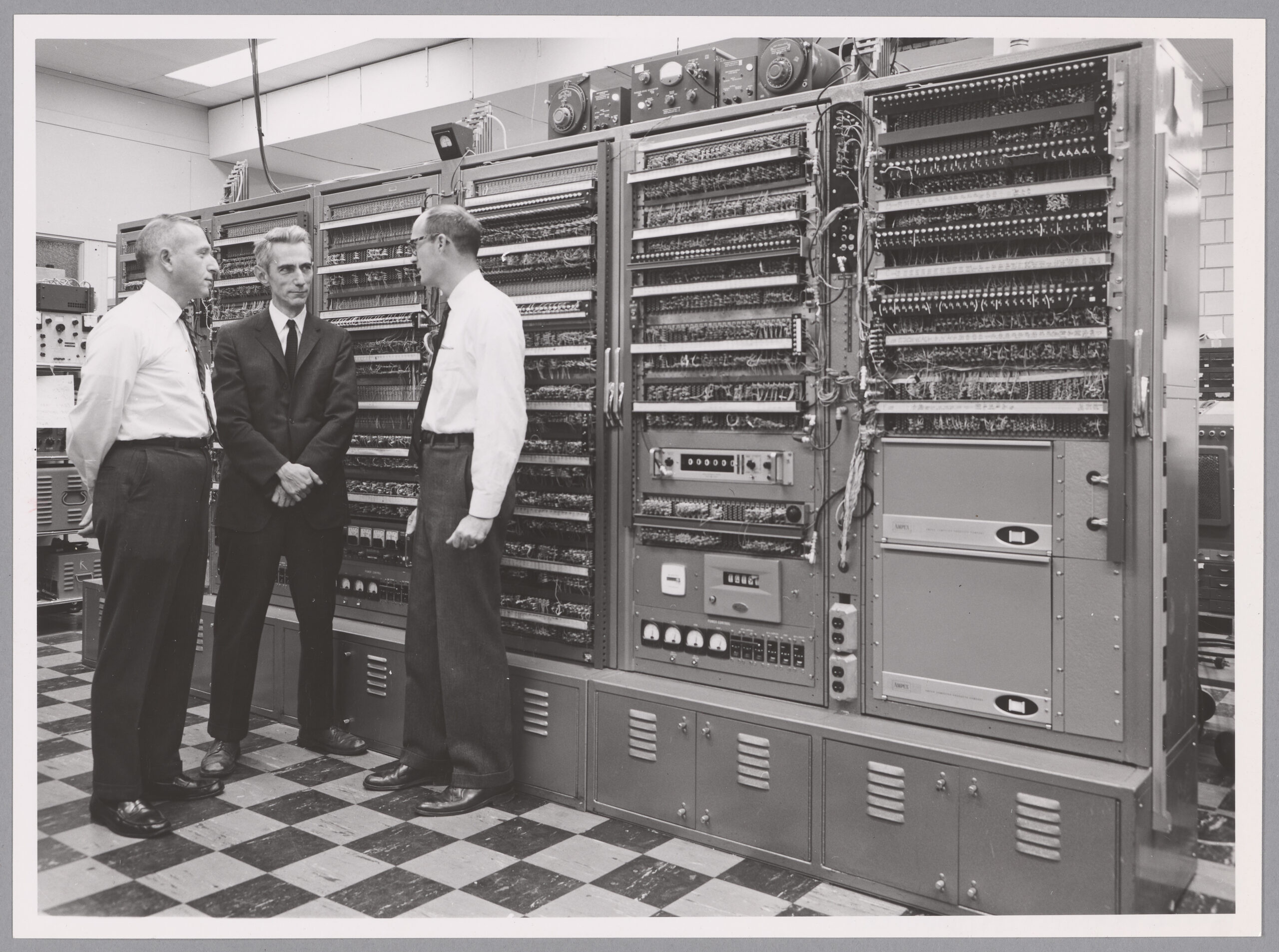Three men stand together in front of a giant analog computer system.