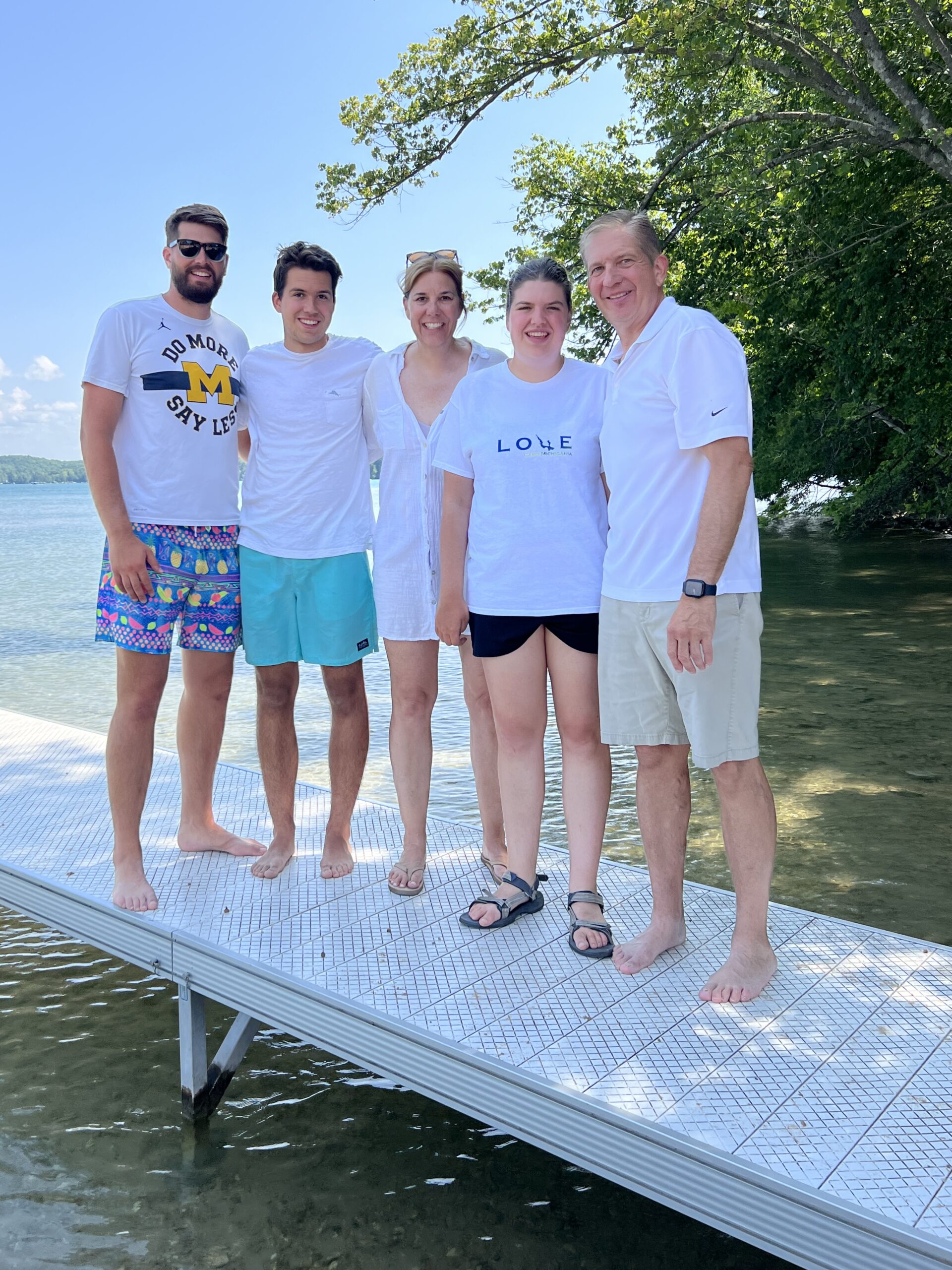 Five people smile on a lakeside dock in casual summer clothes, enjoying Camp Adventure with friends, green trees, and a blue sky.