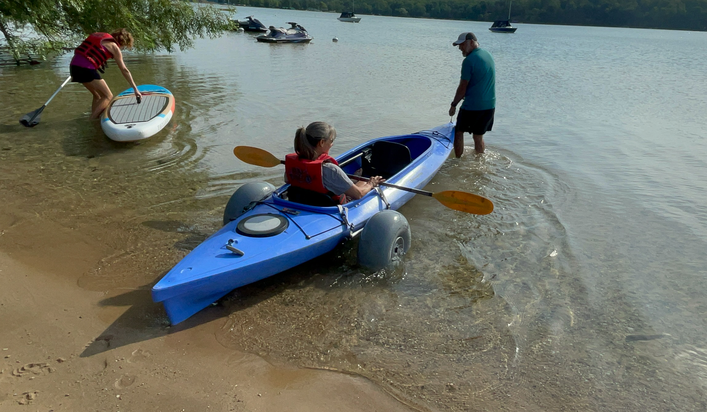 A person in a blue kayak with large wheels is helped into the water as another with a paddleboard stands nearby on a sandy lakeshore.