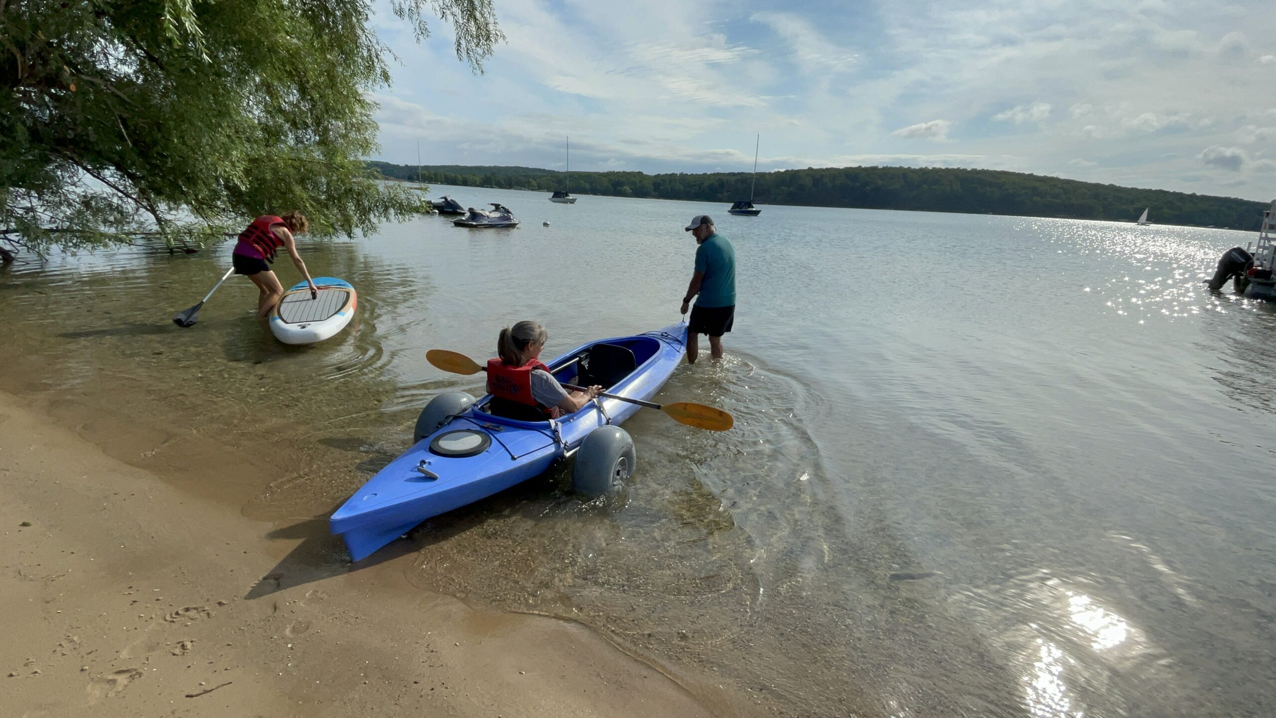 At Camp Michigania, a person in a blue kayak with adaptive wheels is helped into the lake, while another stands by with a paddleboard.
