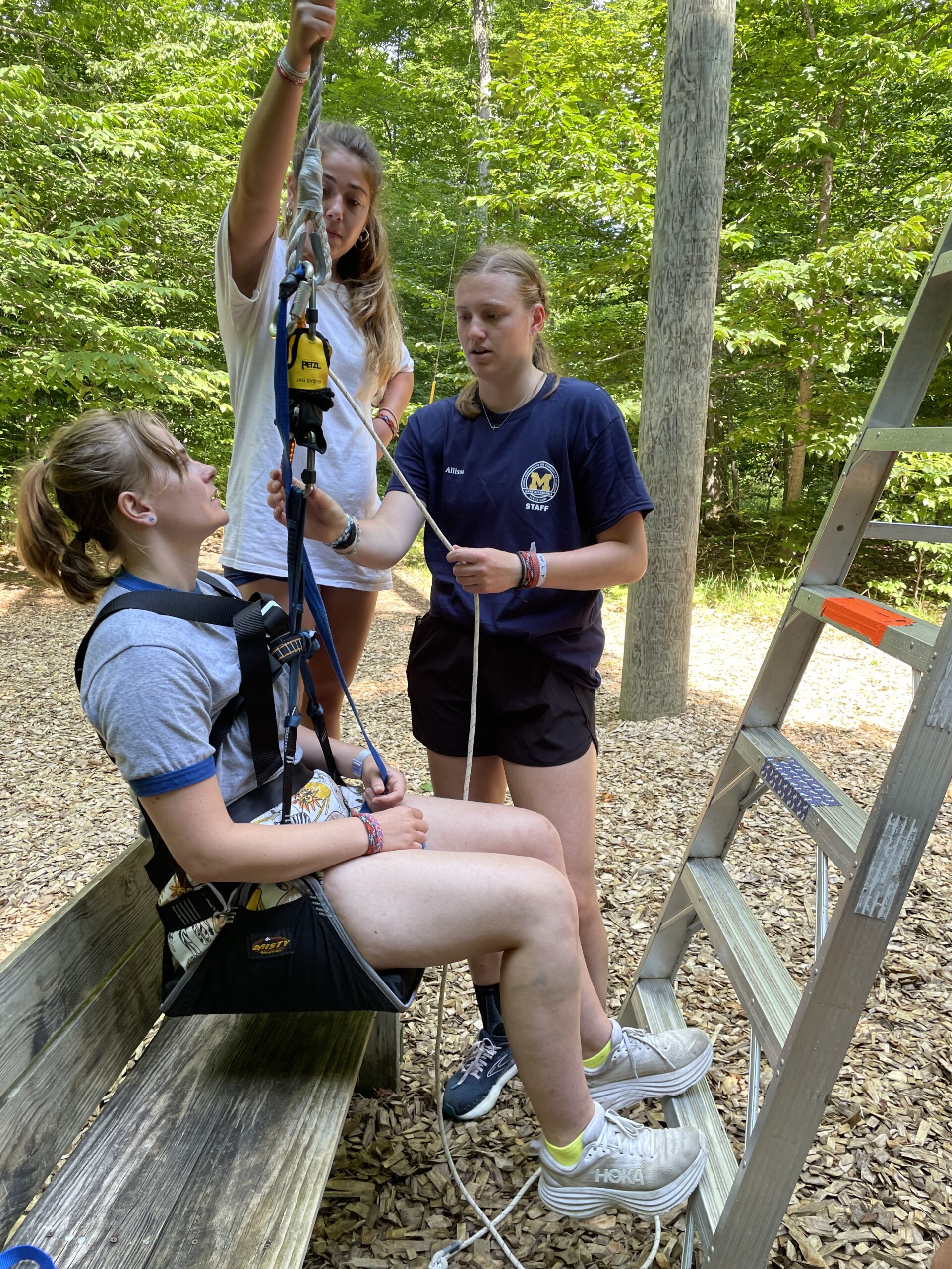 Three young women are by a ladder and bench at Camp; one sits in a harness, while two assist with ropes, amid trees.