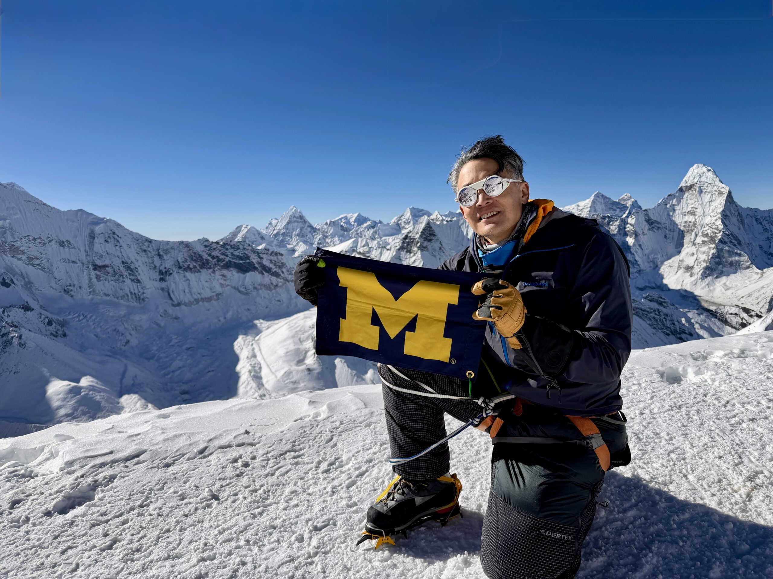 A person in winter climbing gear kneels on a snowy mountain, holding a University of Michigan flag, with mountains and blue sky.