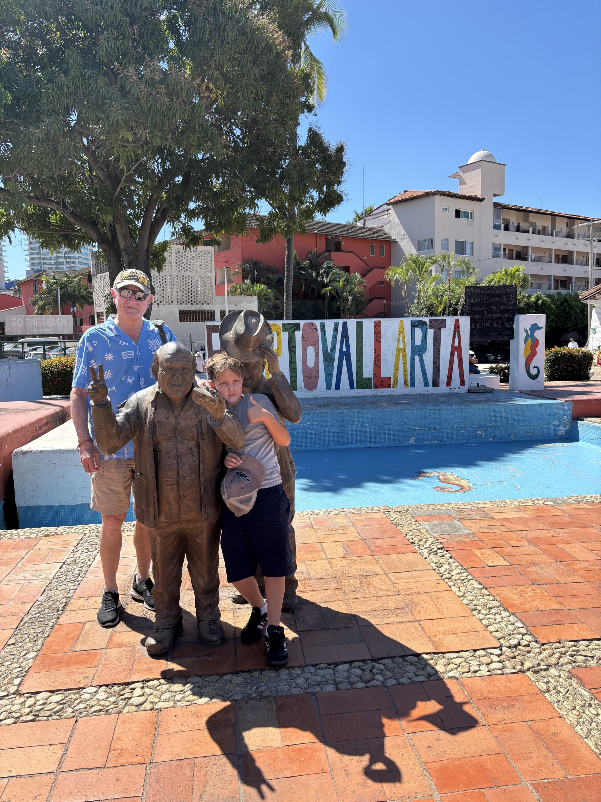 Three people pose with two bronze statues by a colorful “Puerto Vallarta” sign, under a blue sky, with palm trees and buildings.