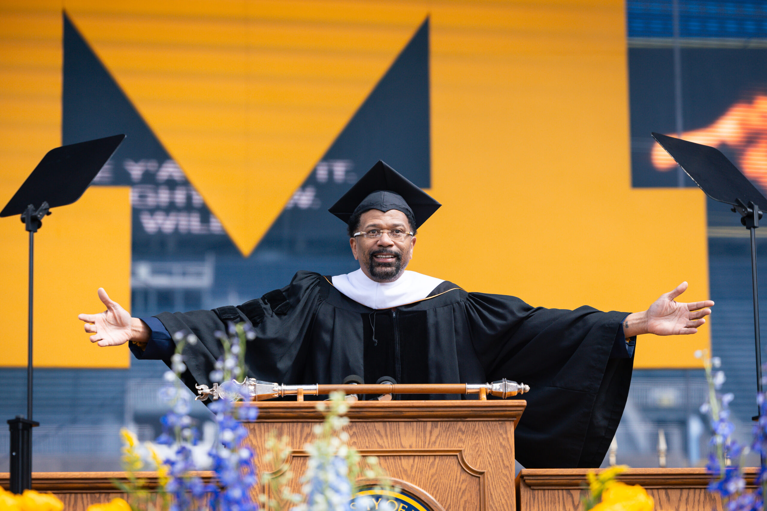 A man in academic regalia stands at a podium with arms outstretched, speaking at an outdoor graduation with yellow, black decor.