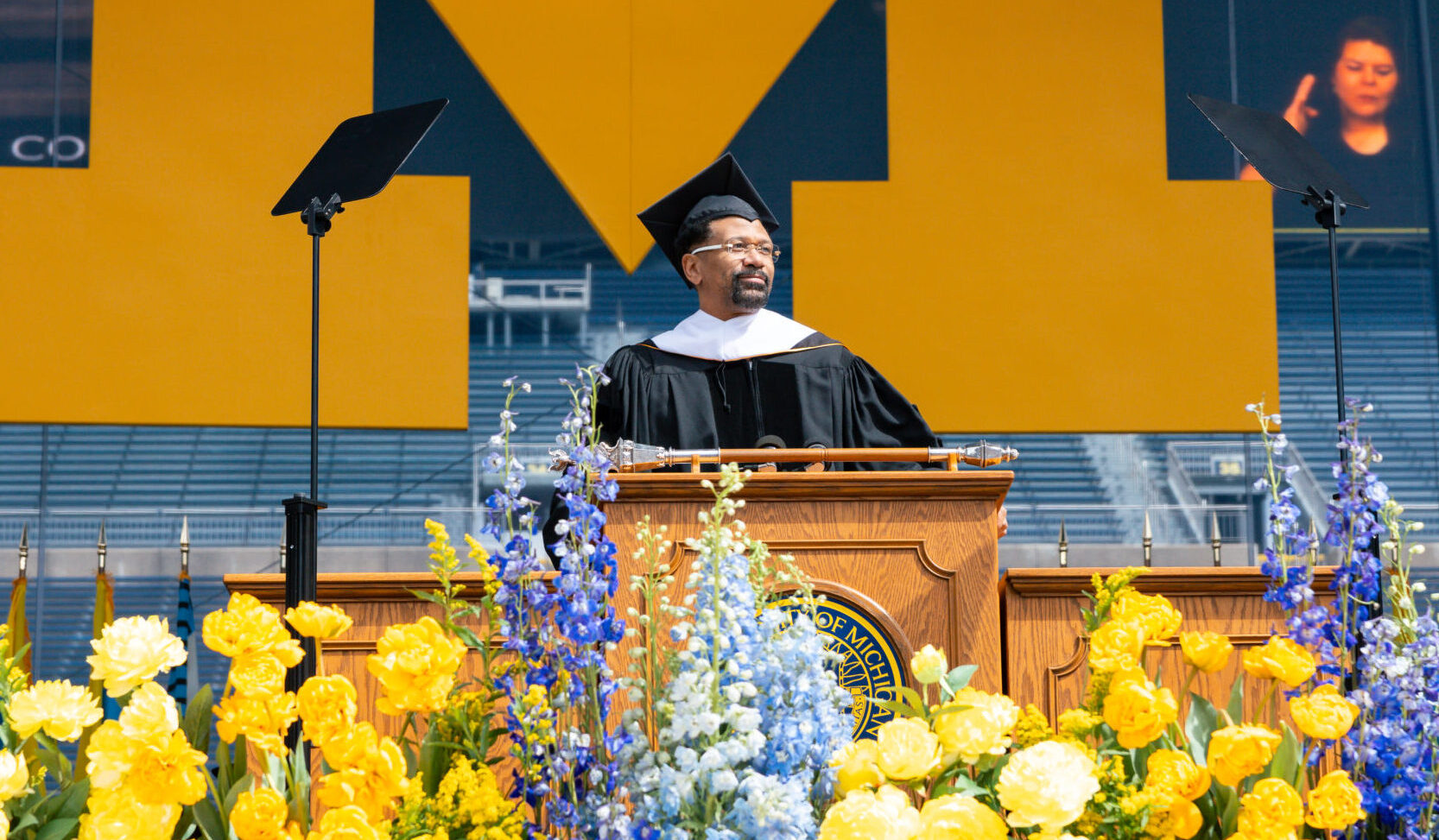 A commencement speaker stands at a podium with yellow, blue flowers, a large yellow M in back, and an interpreter signing at right.