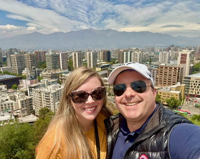 A smiling couple in sunglasses and casual clothes take a selfie with a blue cityscape, tall mountains, and a partly cloudy sky behind.