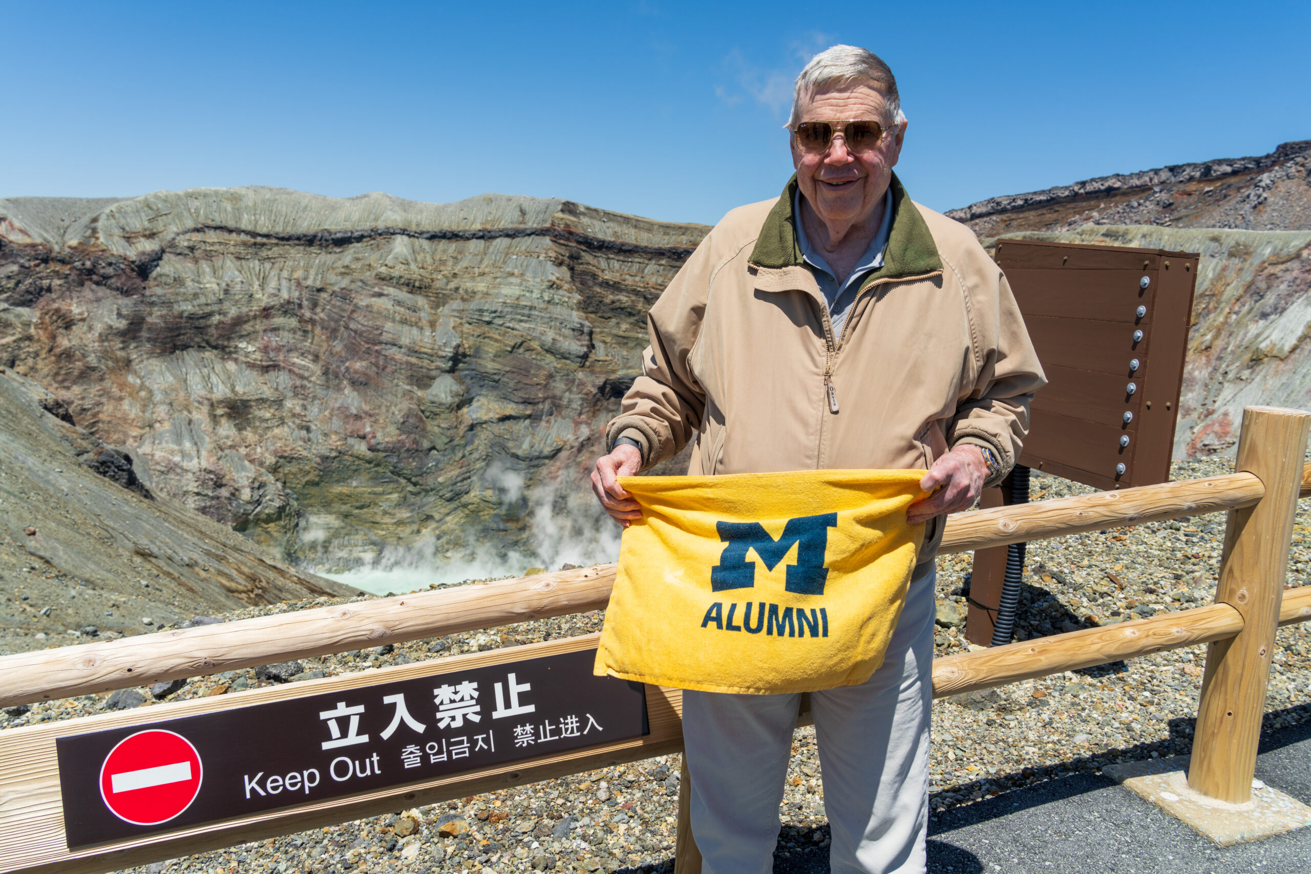 An older man in sunglasses smiles and holds a yellow M Alumni towel by a Keep Out sign, with blue sky, rocky crater behind him.