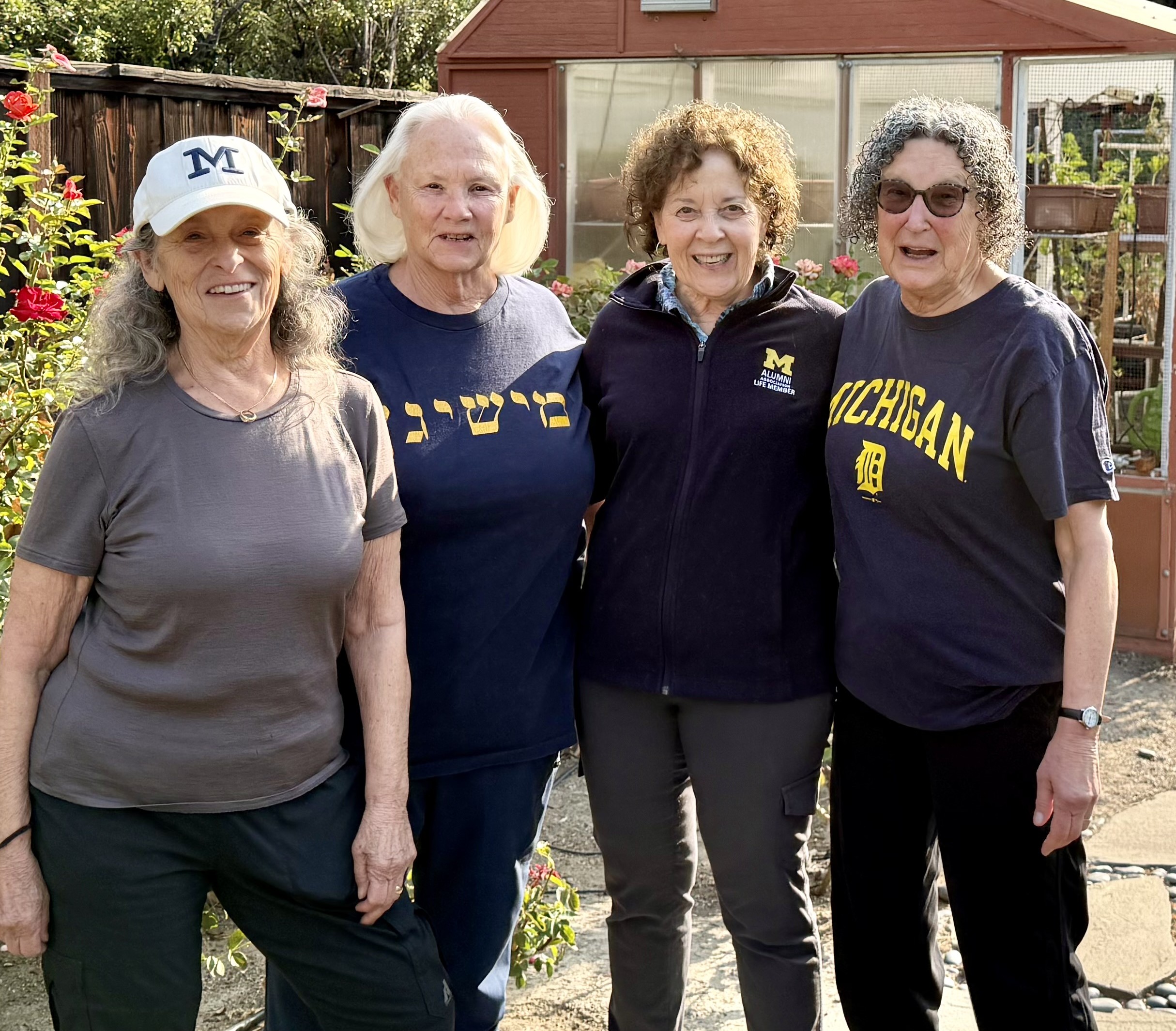 Four older women stand smiling outdoors, arm in arm. Three wear Michigan Blue, and one wears a shirt with Hebrew writing.