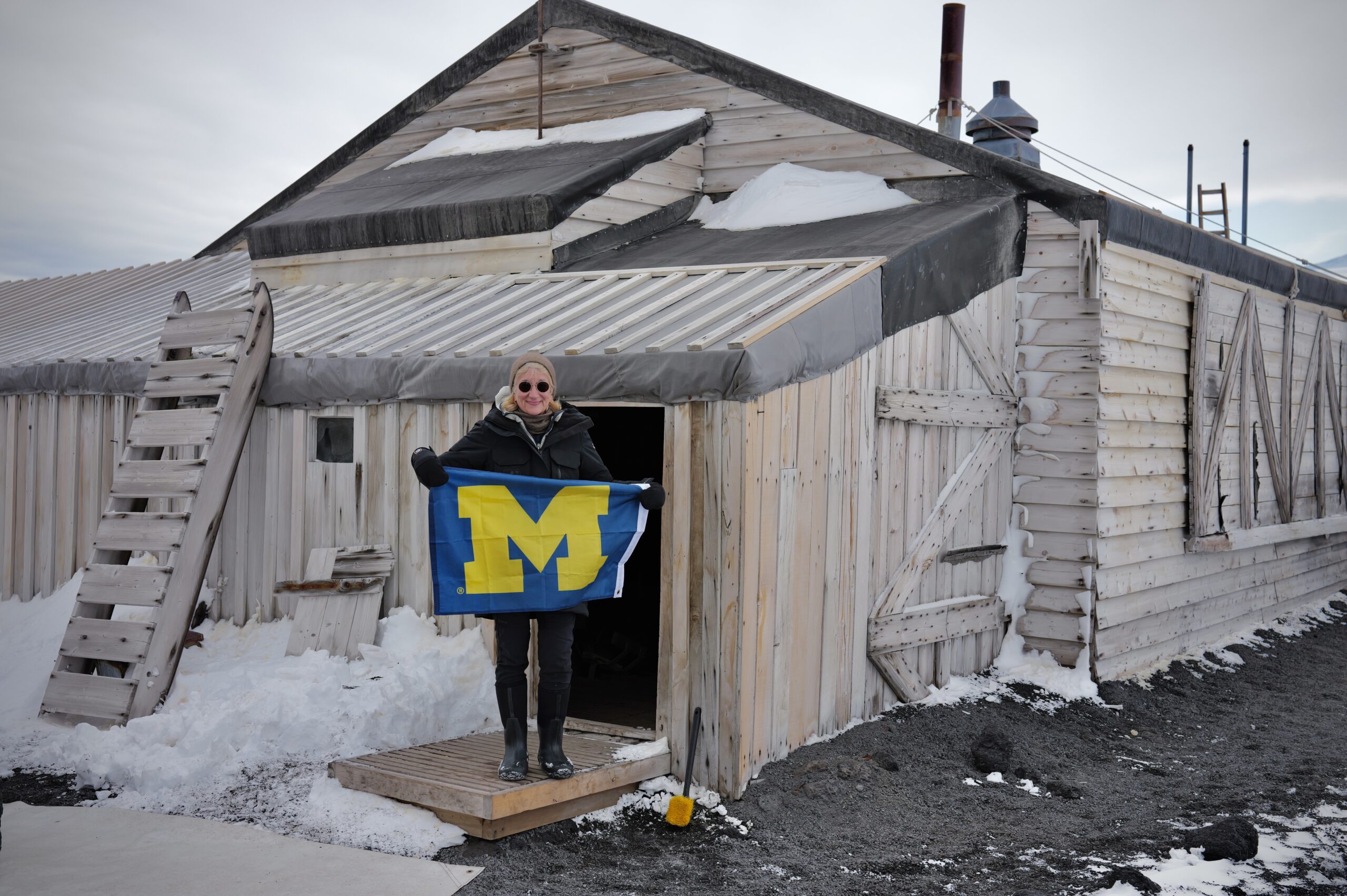 A person stands outside a weathered wooden building in snow and ice, holding a blue flag with a large yellow M.
