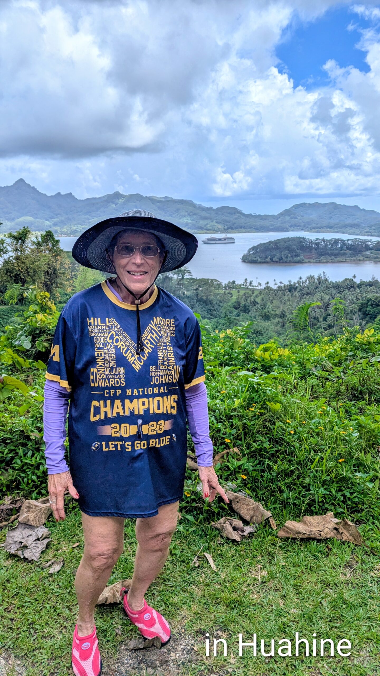 An older woman in a blue sports shirt, sunhat, and pink sandals smiles on a hilltop with Huahine island behind her. Text: "in Huahine – Summer 2026.