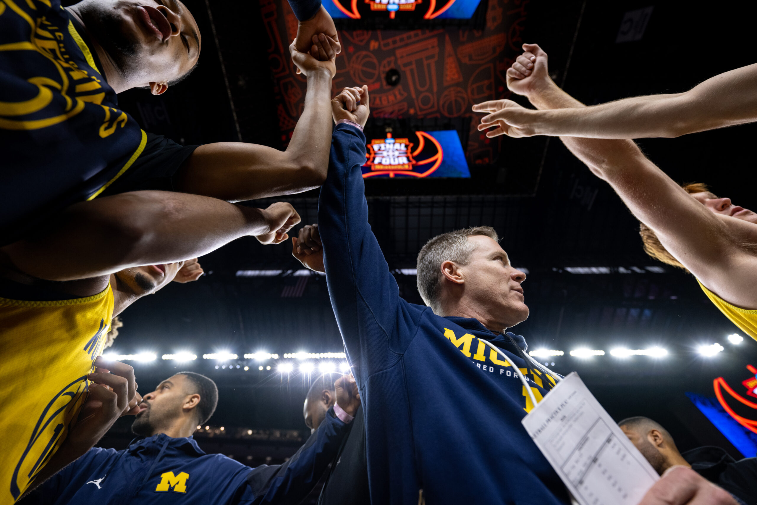 The men's basketball team gathers around the coach to put their hands in the middle.