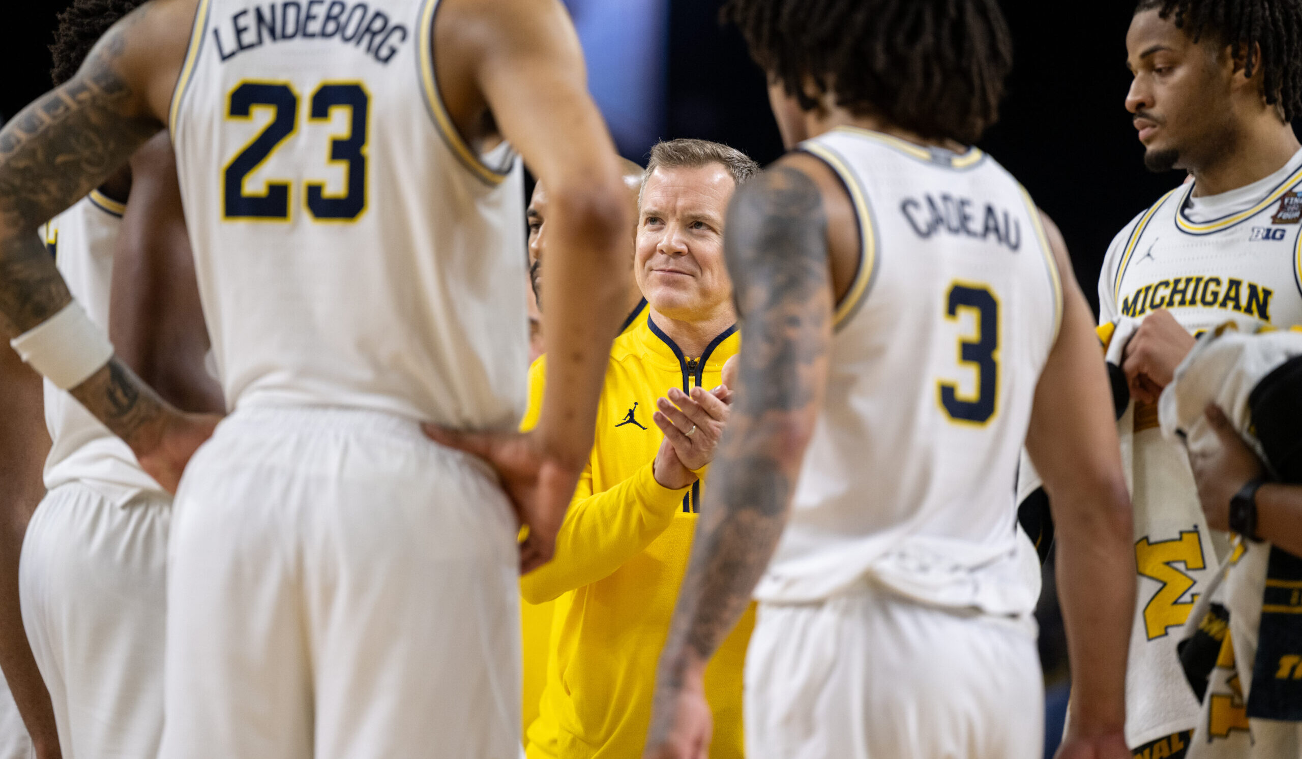 A basketball coach in a yellow jacket addresses his white-uniformed Michigan team in a huddle, delivering a Built to Win message.
