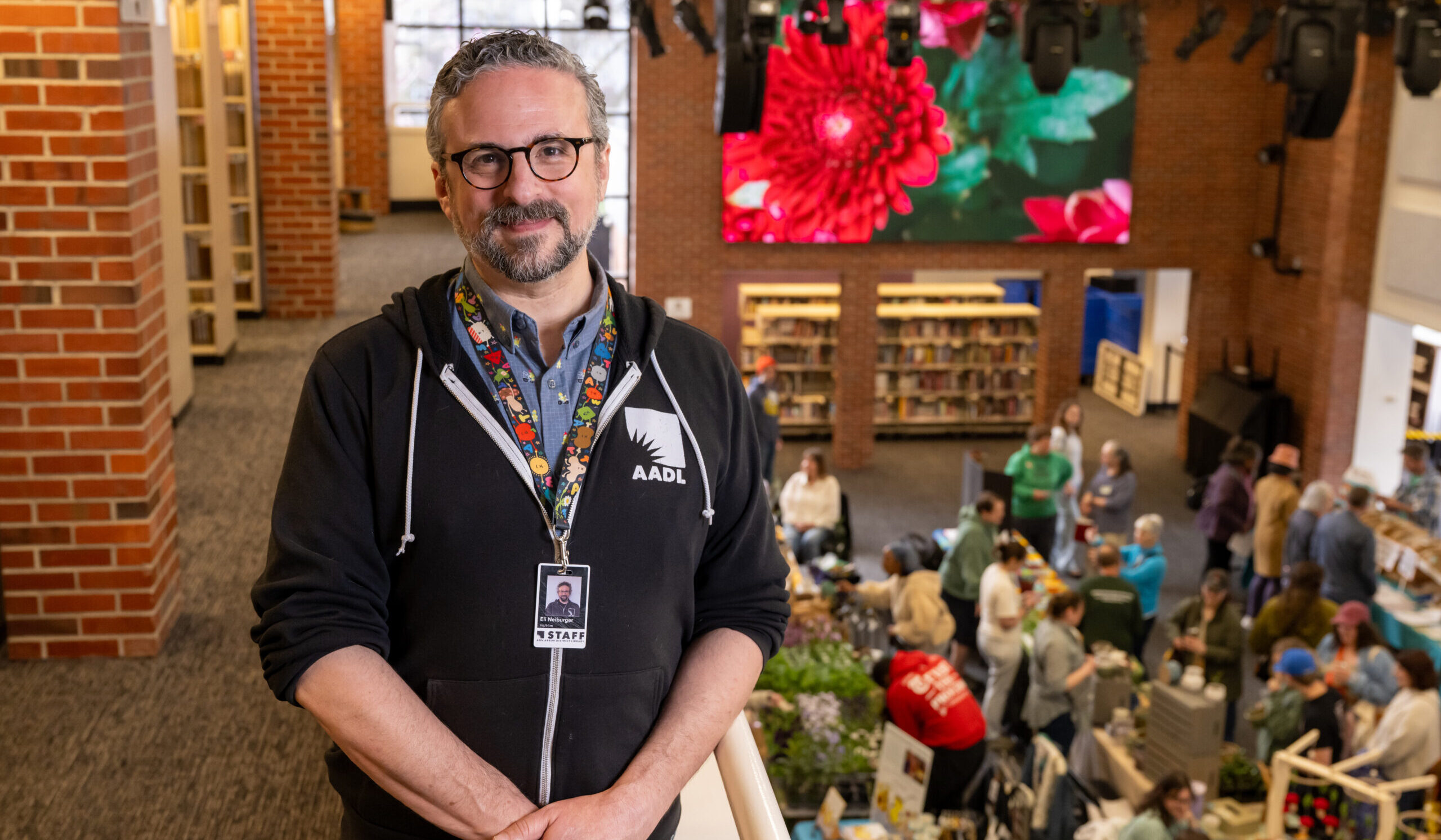 Eli Neiburger, wearing glasses and an AADL badge, stands on a balcony at Ann Arbor library, overlooking a busy event and digital flower display.