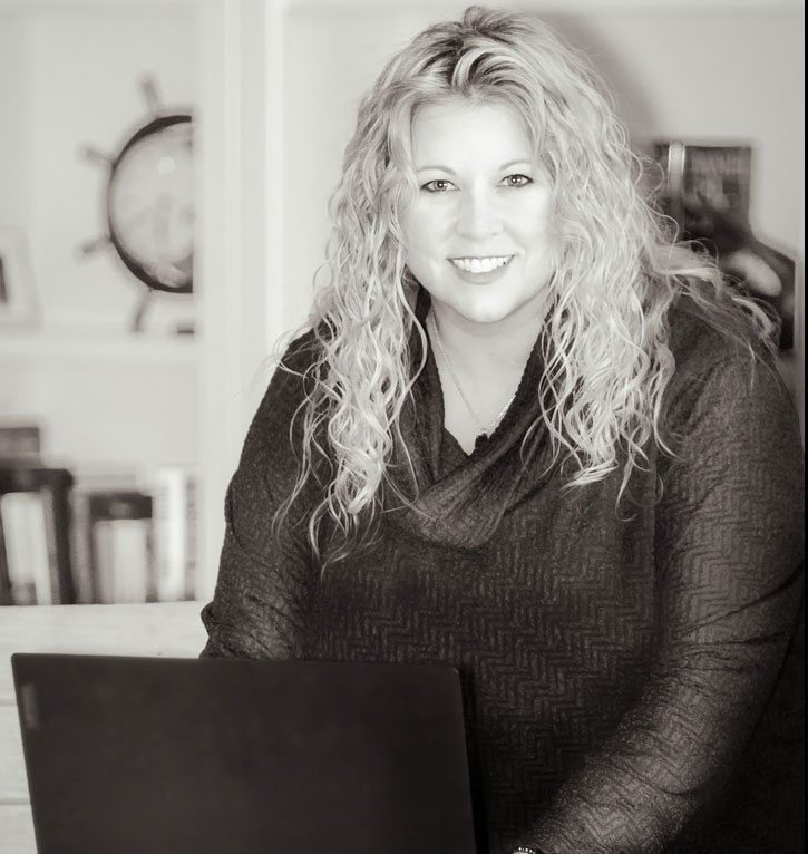A person with long, curly hair smiles at a desk behind a laptop.