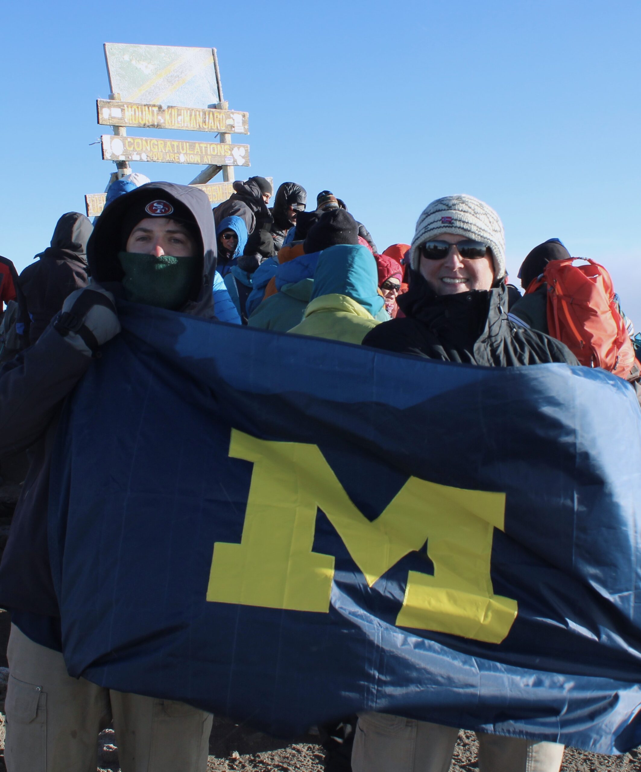 Two people in winter clothing hold a blue flag with a yellow M at Mount Kilimanjaro’s summit.