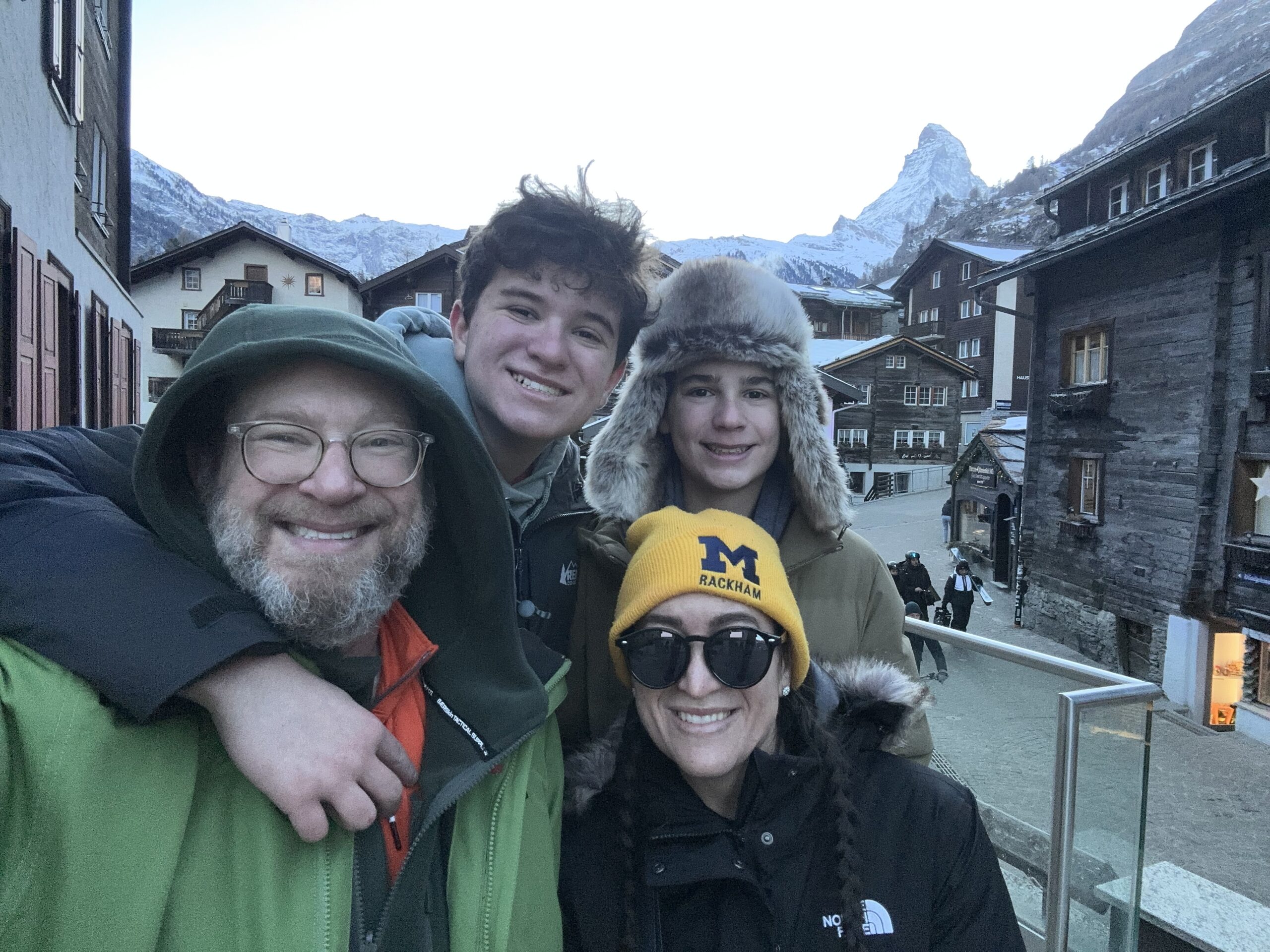 Four people in winter clothing smile outdoors in a mountain village, with snowy peaks and wooden buildings behind them.