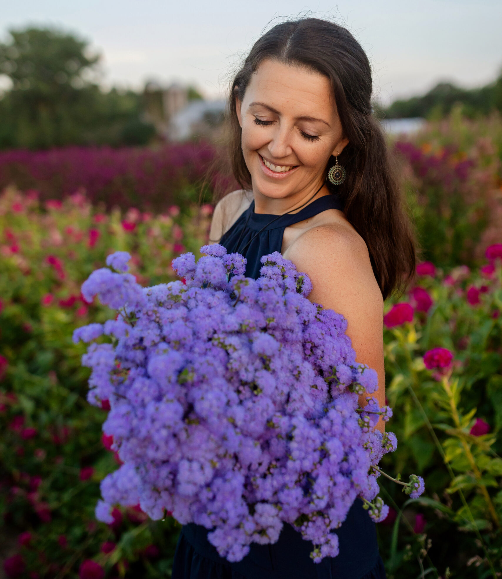 A woman with long dark hair in a navy dress smiles, holding purple flowers, in a colorful field.