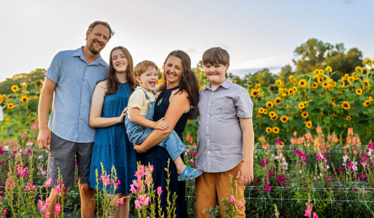 A family of five—two adults, three children—smile in a field of sunflowers and blooms as they pose for a sunny day photo.