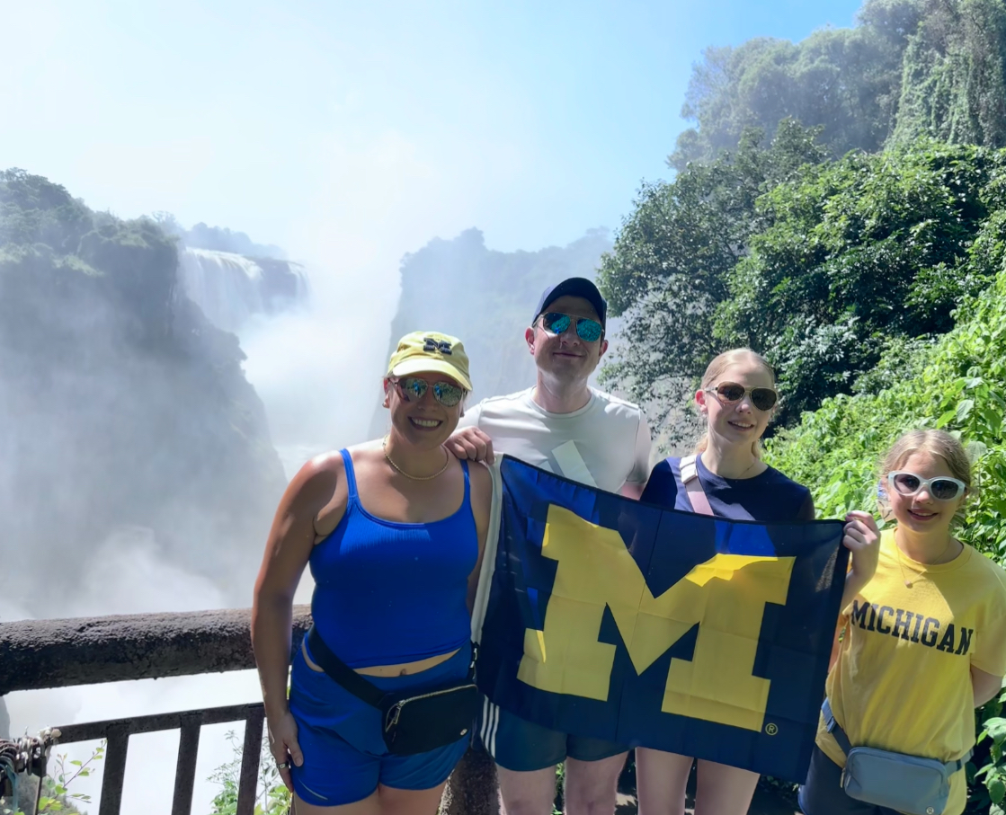 Four people in Michigan gear smile by a wooden railing, holding a blue and yellow "M" flag near a misty waterfall and greenery.