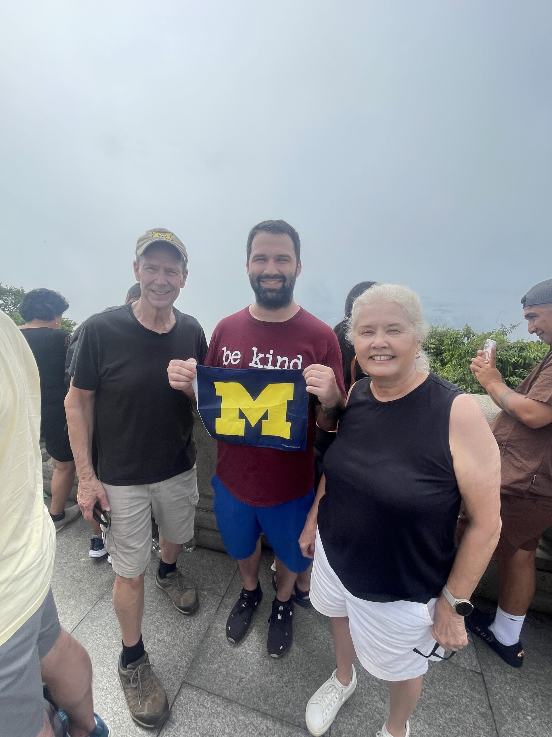 Three people pose outdoors, smiling. The middle person holds a blue University of Michigan flag and wears a "be kind" shirt.