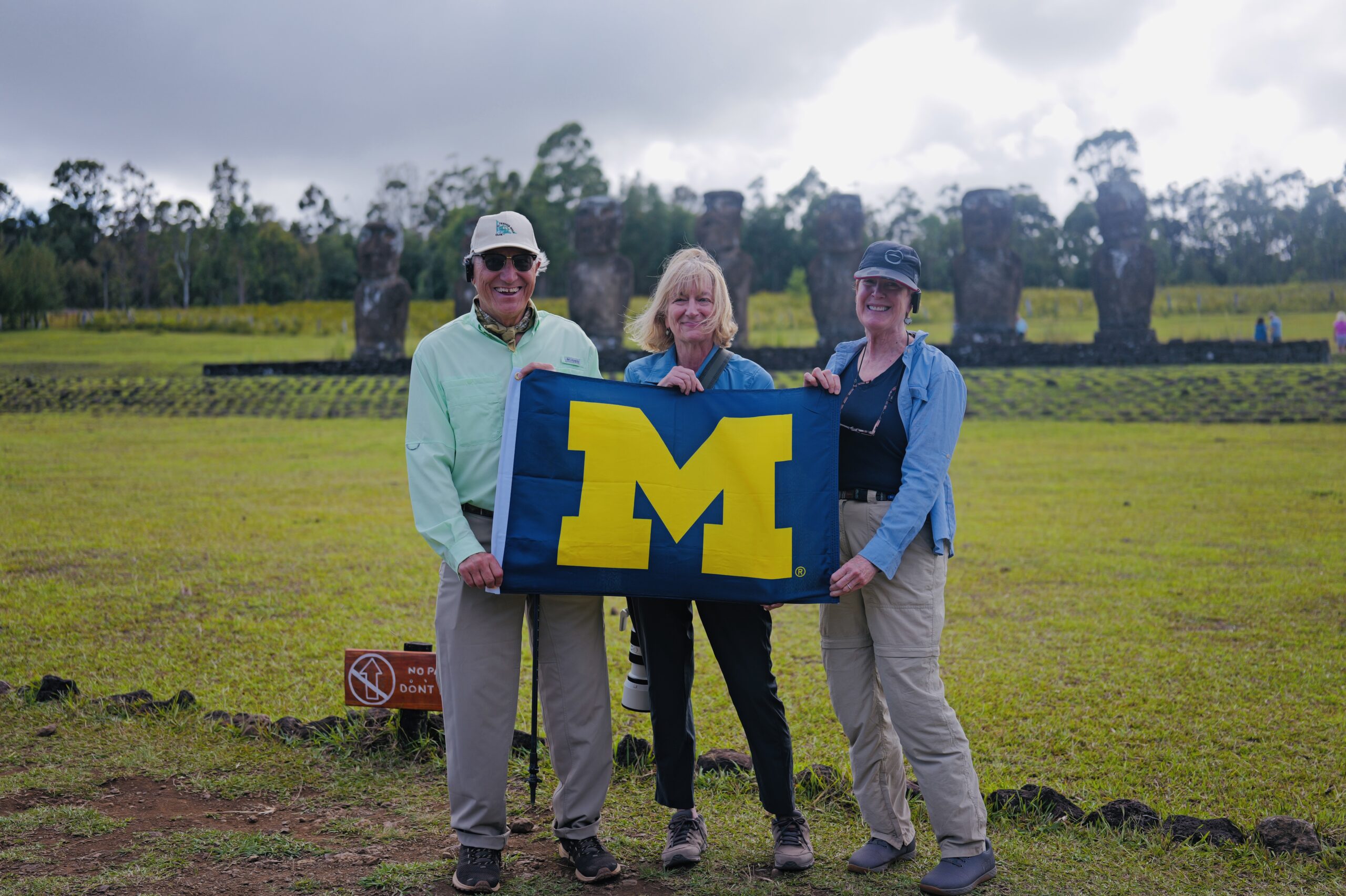 Three people stand on grass holding a blue University of Michigan flag, with Moai statues in back, under cloudy skies.