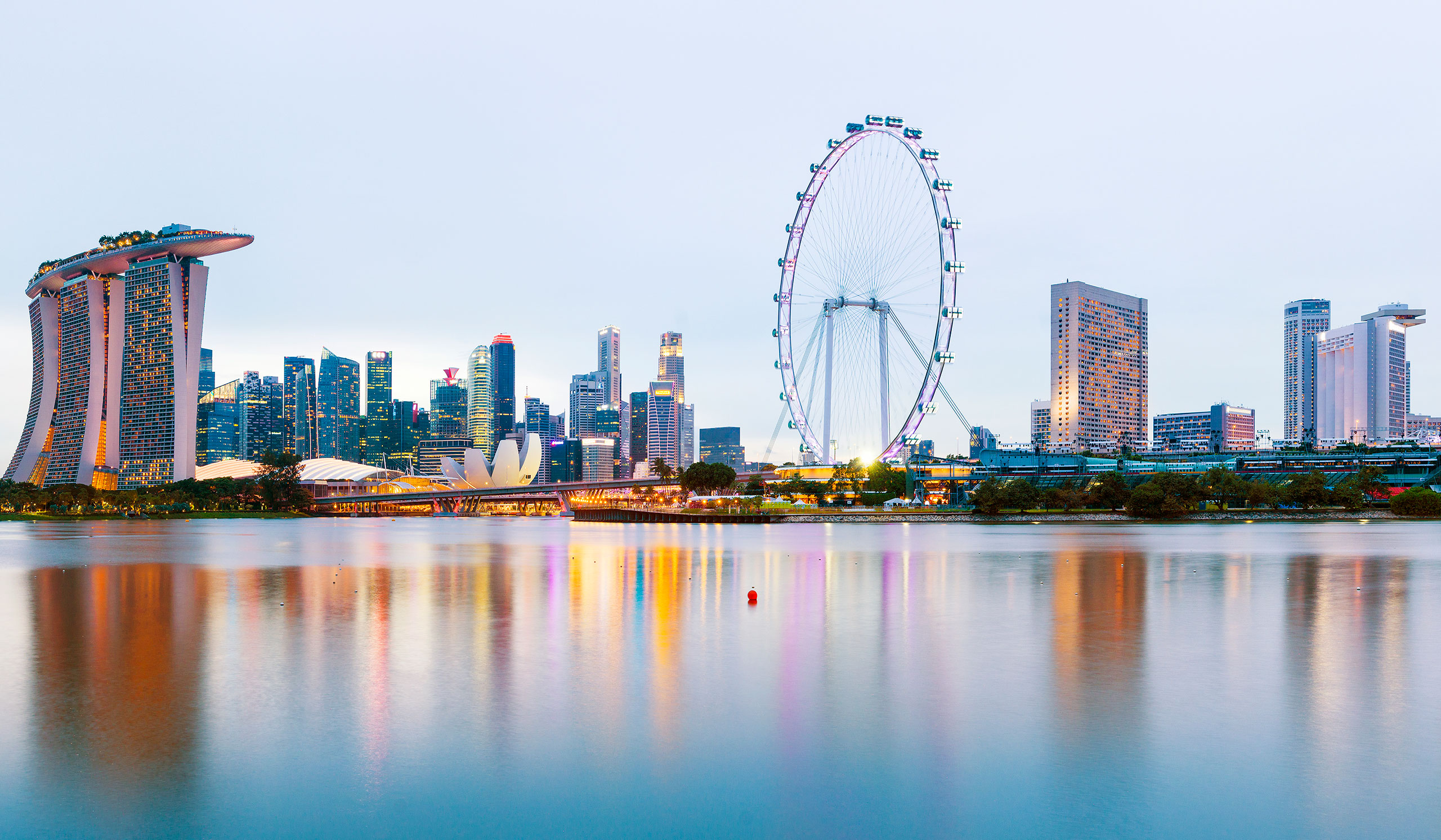 Singapore skyline at dusk, with Marina Bay Sands hotel, the Singapore Flyer, and modern buildings reflected on the calm bay.