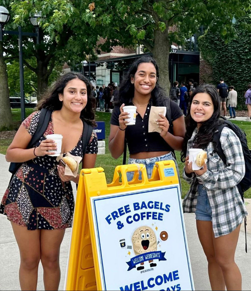 Three smiling students hold bagels and drinks near a sign for Welcome Wednesdays in front of the Alumni Center.