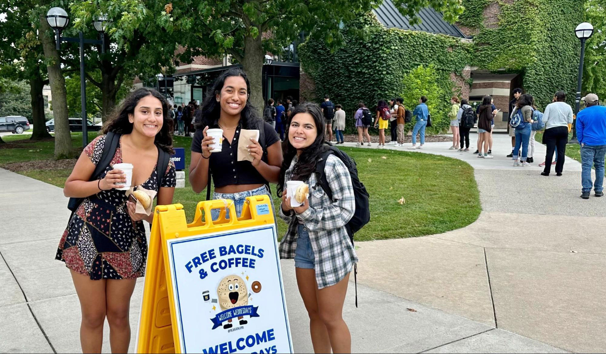 Three smiling students hold bagels and drinks near a sign for Welcome Wednesdays in front of the Alumni Center.