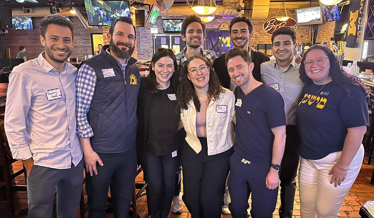 Nine people wearing name tags smile together in a bar with University of Michigan flags in the background.